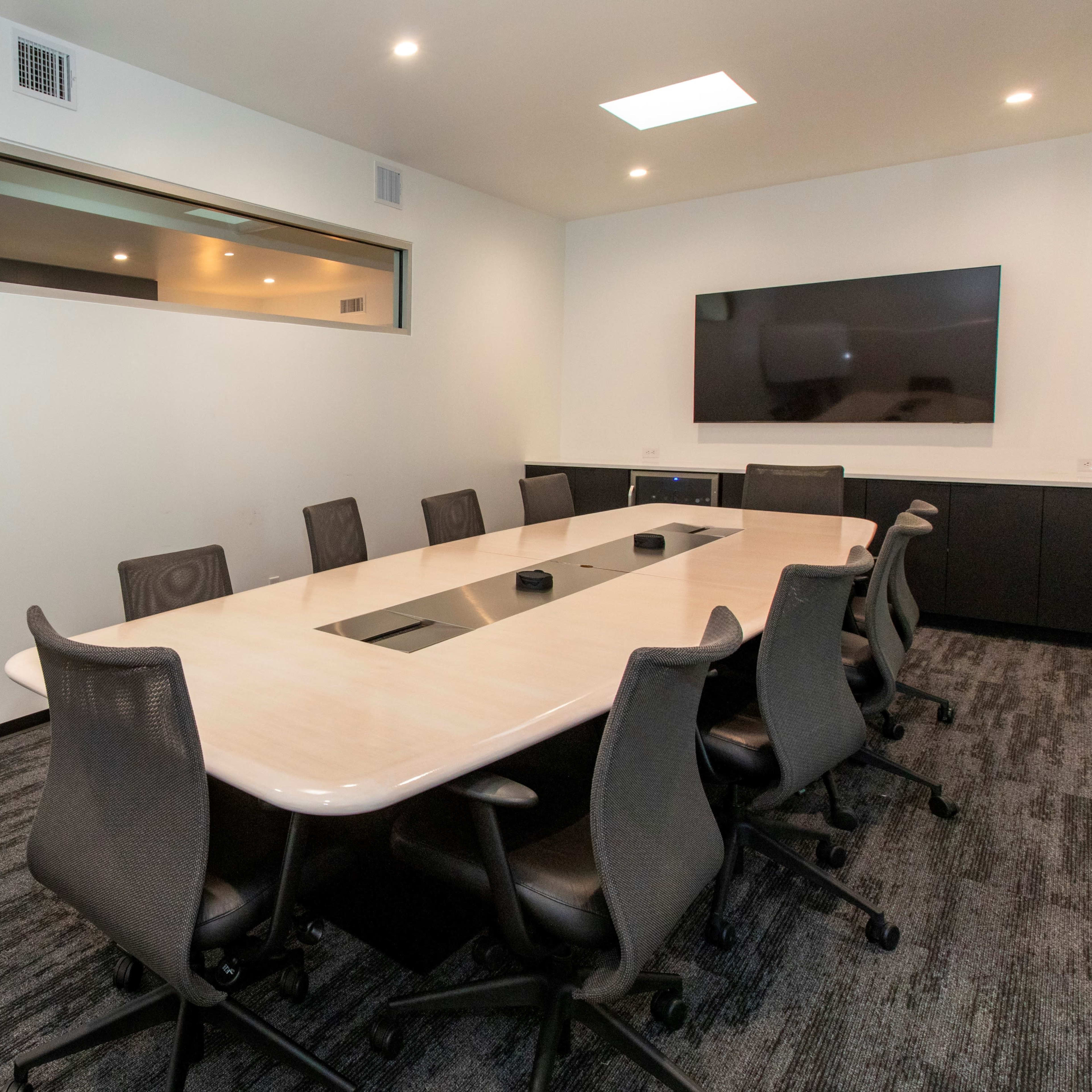 A modern conference room features a long oval table surrounded by multiple black ergonomic chairs, with a wall-mounted TV and a window providing natural light.