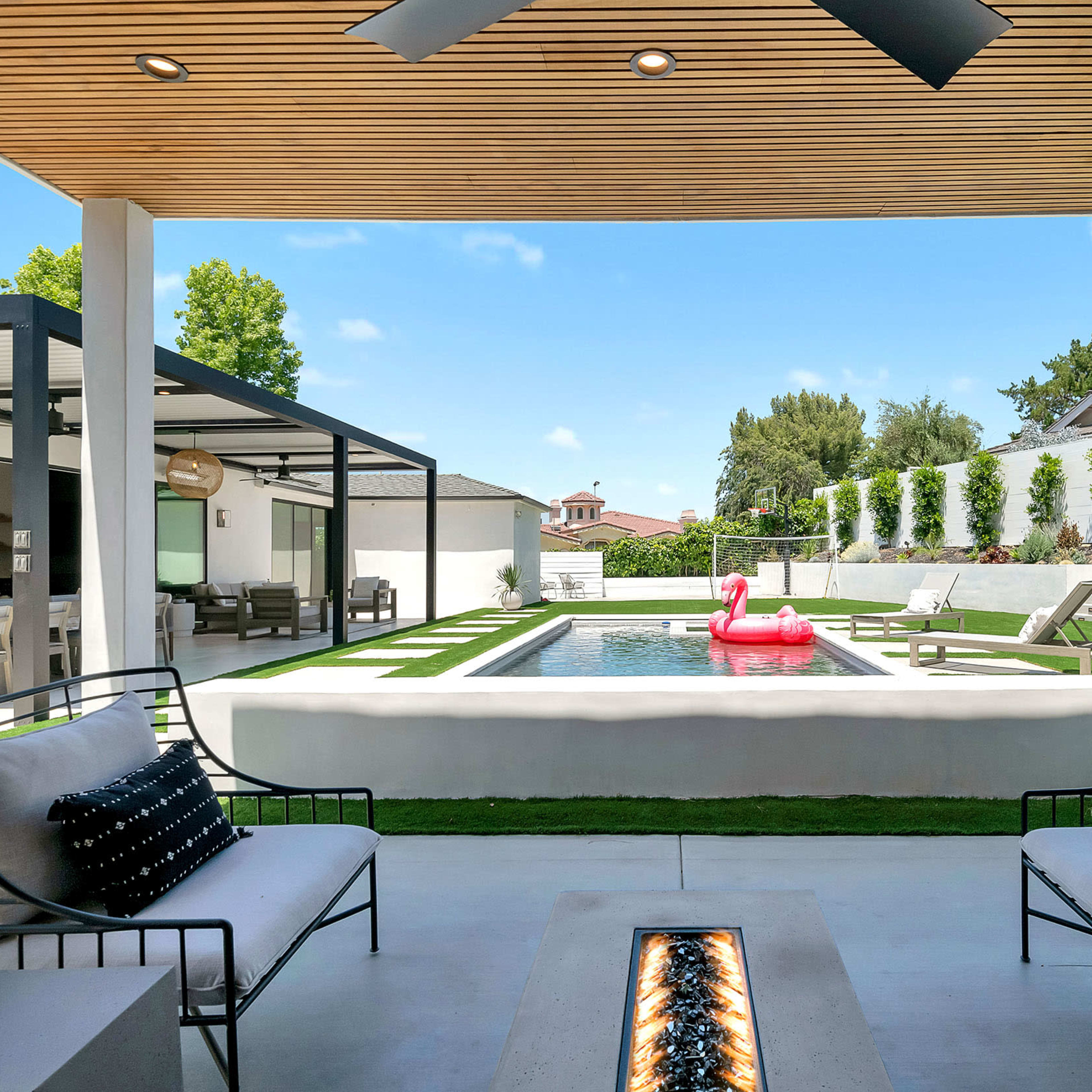 A modern outdoor living space features a rectangular pool with a pink inflatable flamingo, surrounded by seating areas and a landscaped yard.
