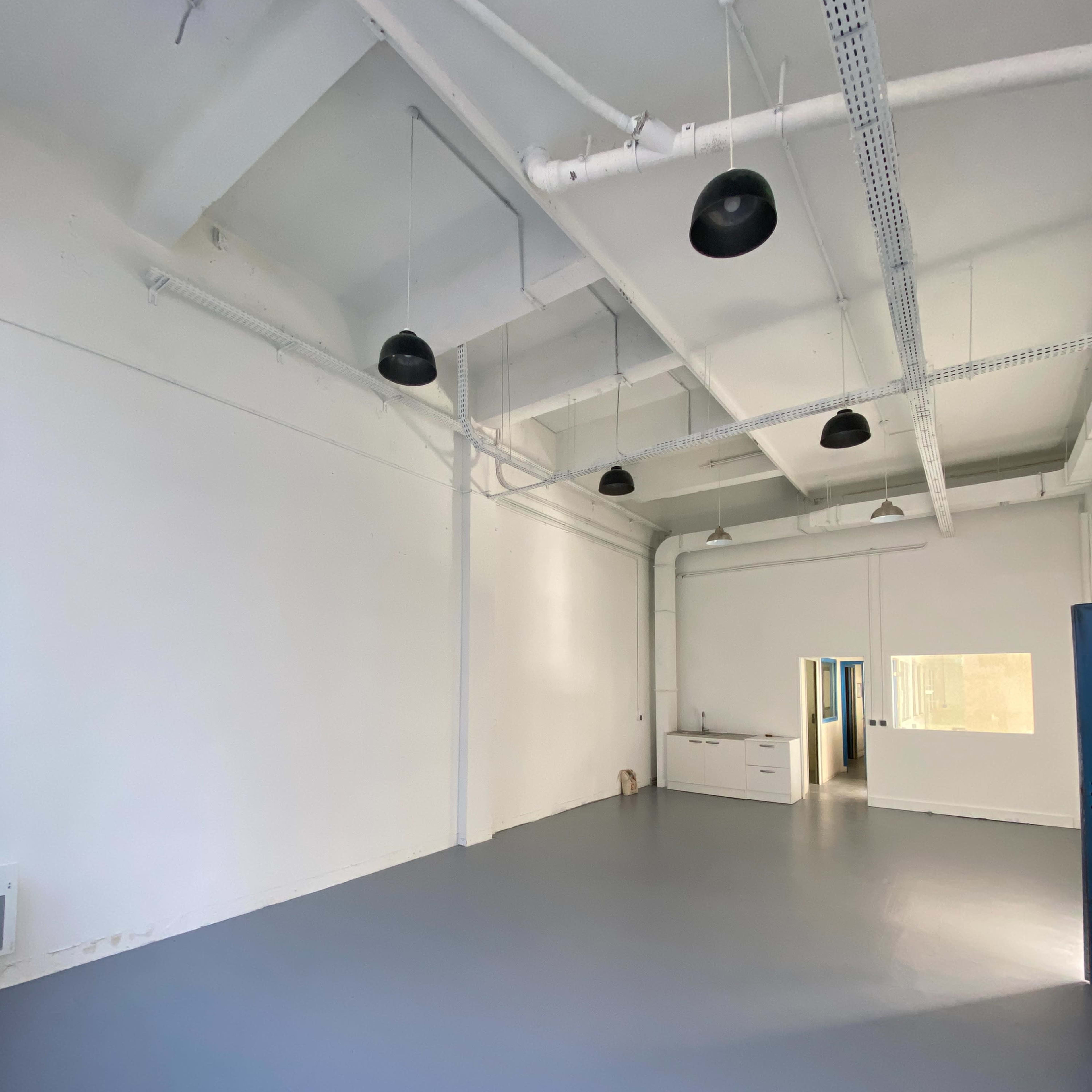A spacious, empty room with white walls and gray flooring, featuring several black ceiling lights and a small kitchenette in the background.
