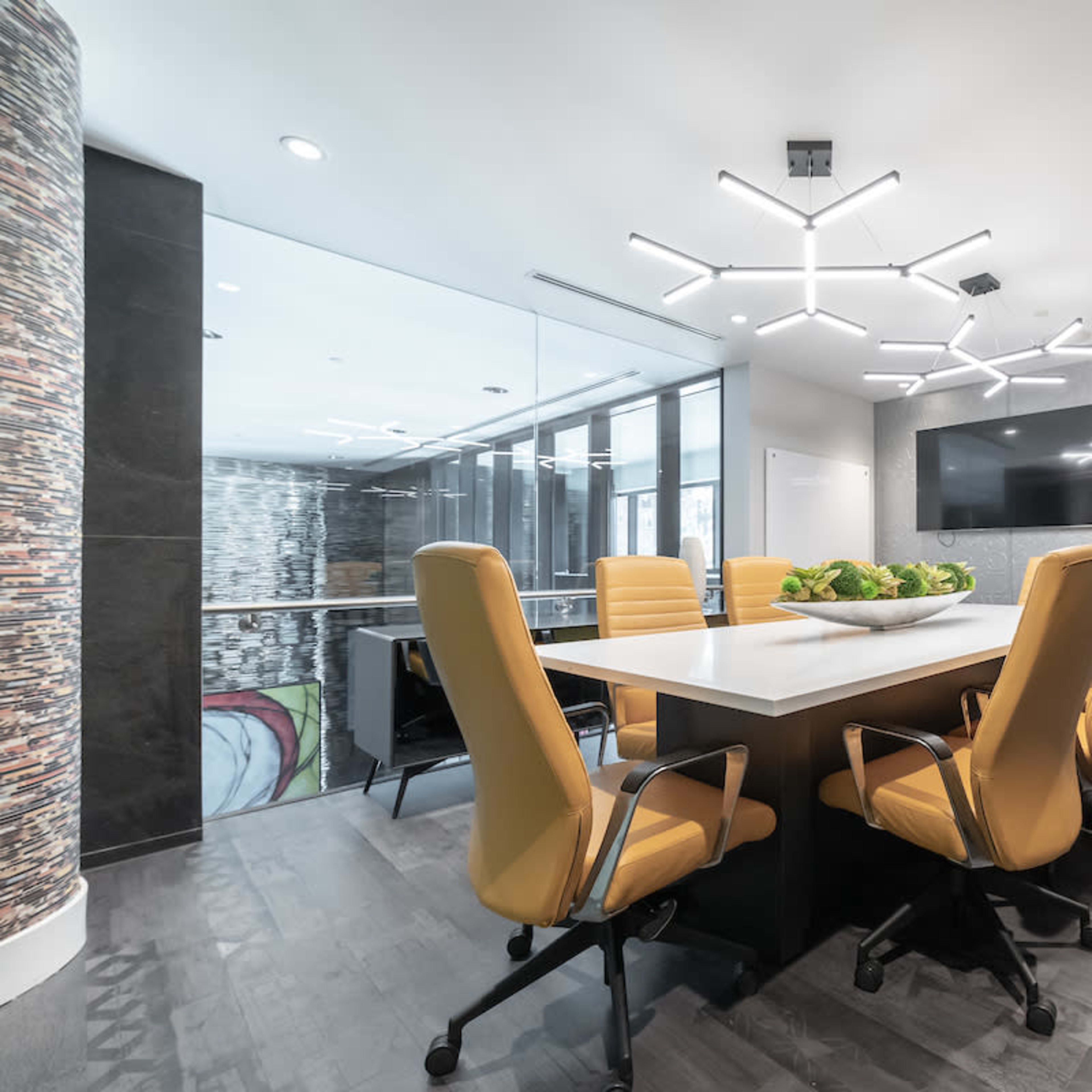 A modern conference room features a large white table surrounded by yellow chairs, with a television and decorative elements in the background.