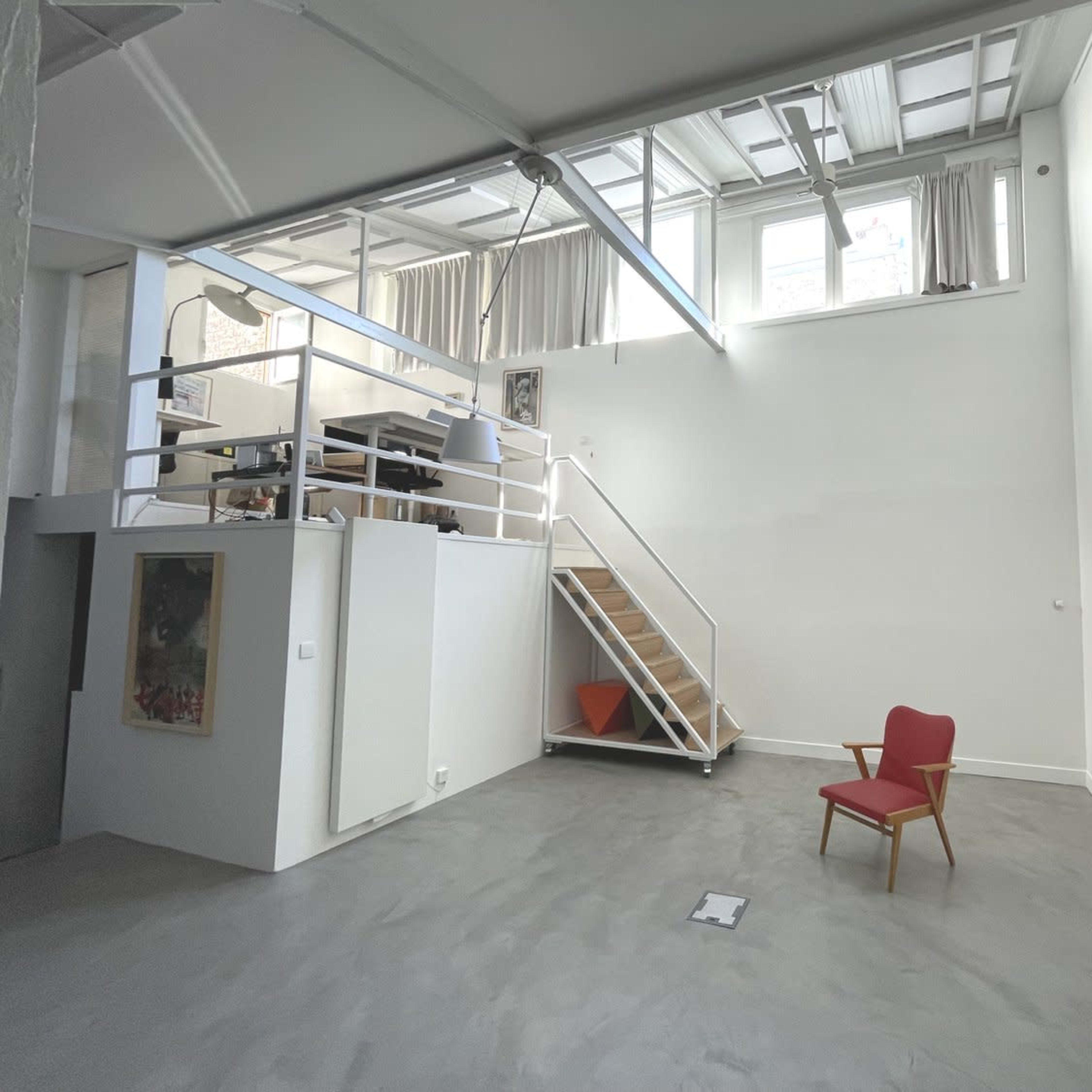A spacious room with a wooden staircase leading to a loft area, a red chair on the floor, and large windows along the upper portion of the wall.