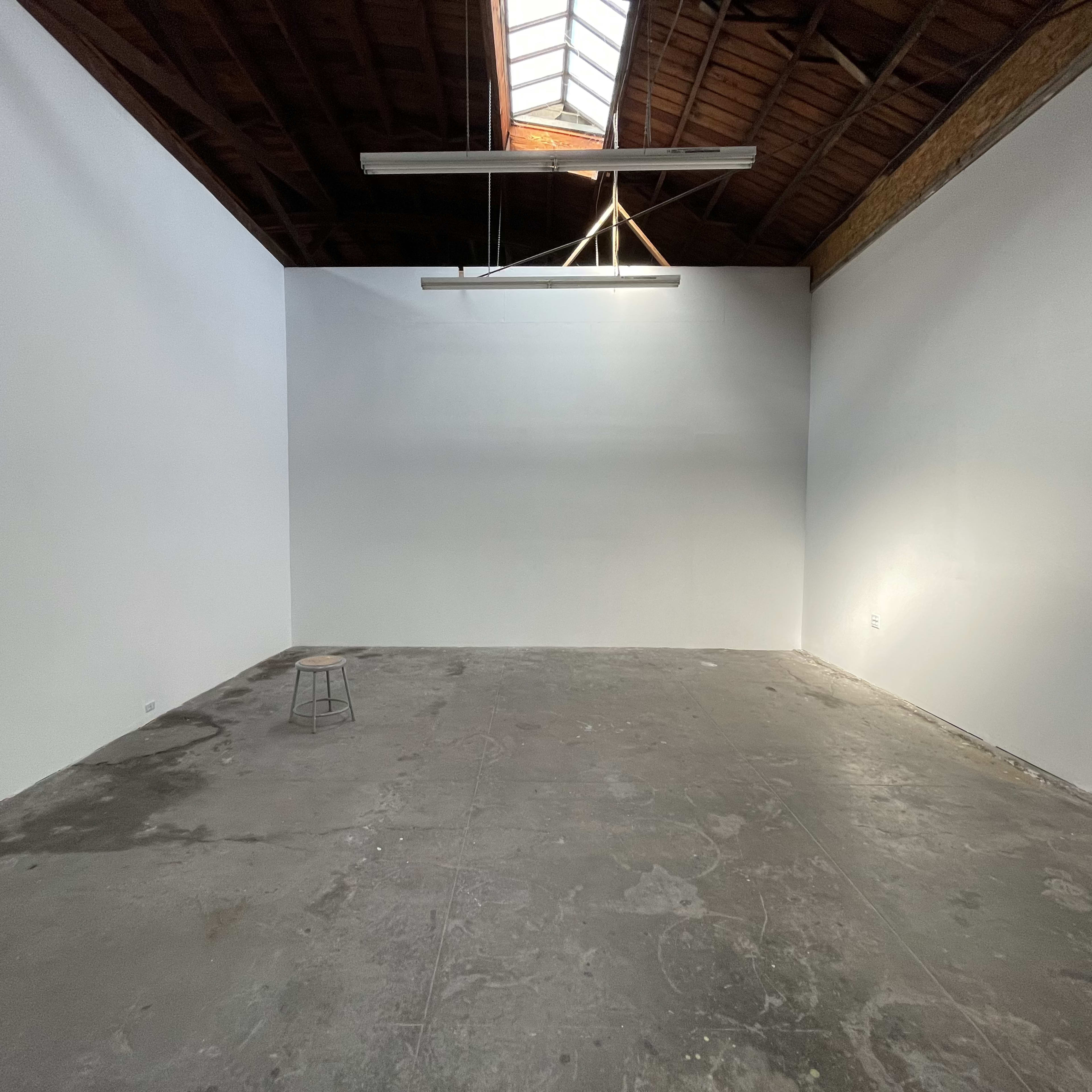 An empty room with white walls, a concrete floor, and a single stool under a skylight.