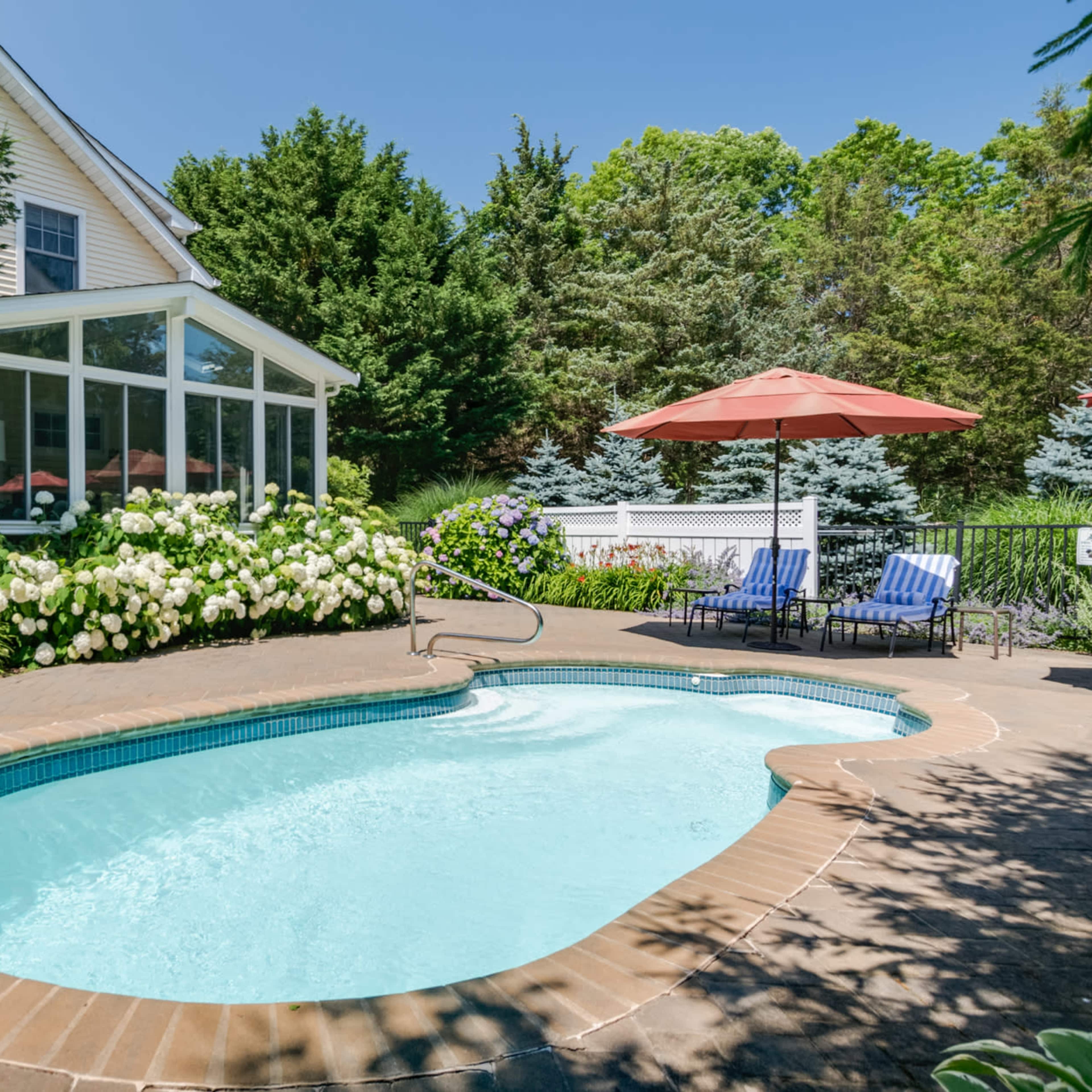 A small, in-ground swimming pool is surrounded by a stone patio, outdoor seating, and landscaped gardens with flowering plants.