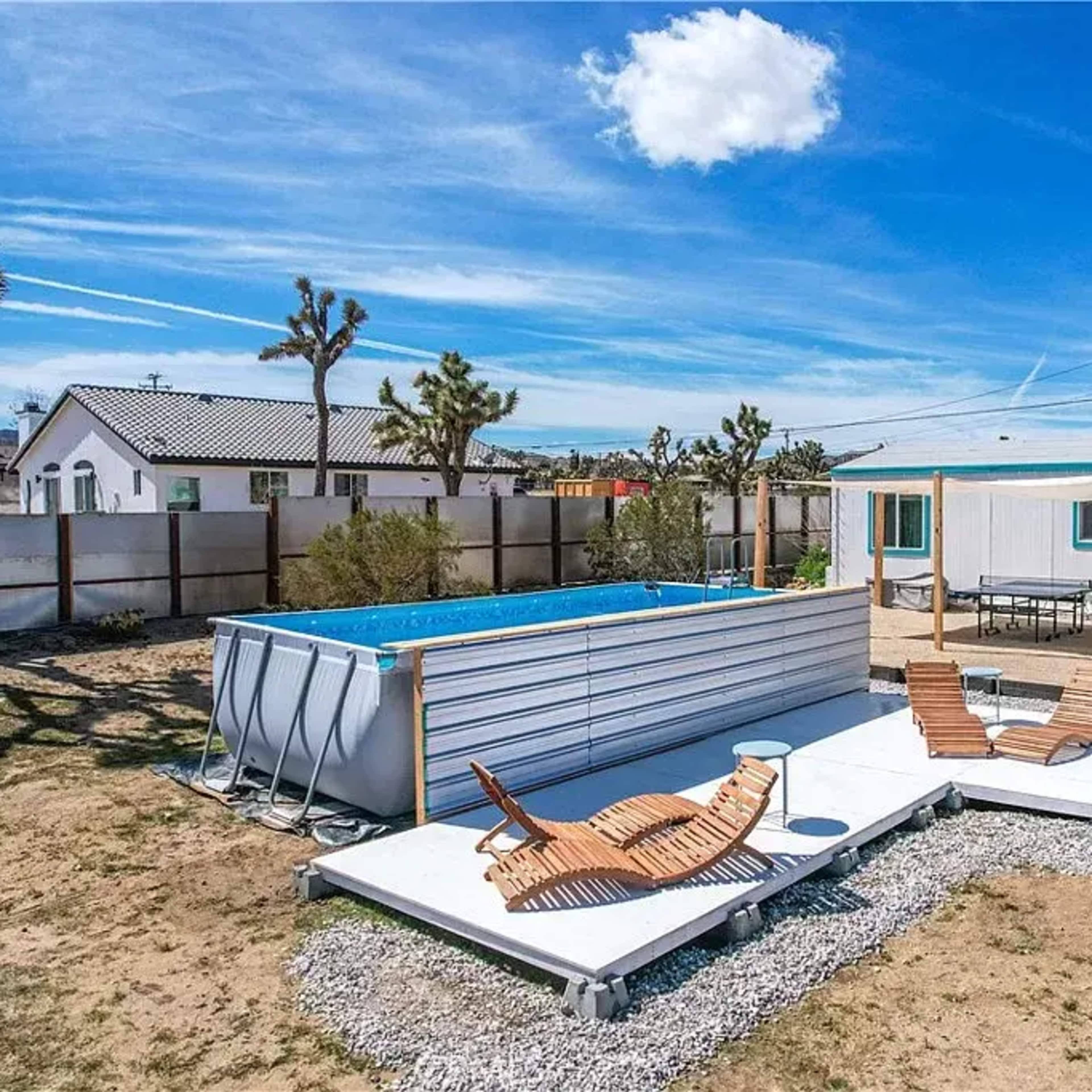 A backyard with a above-ground pool, wooden lounge chairs, and a modern house featuring a cactus mural in a desert setting.
