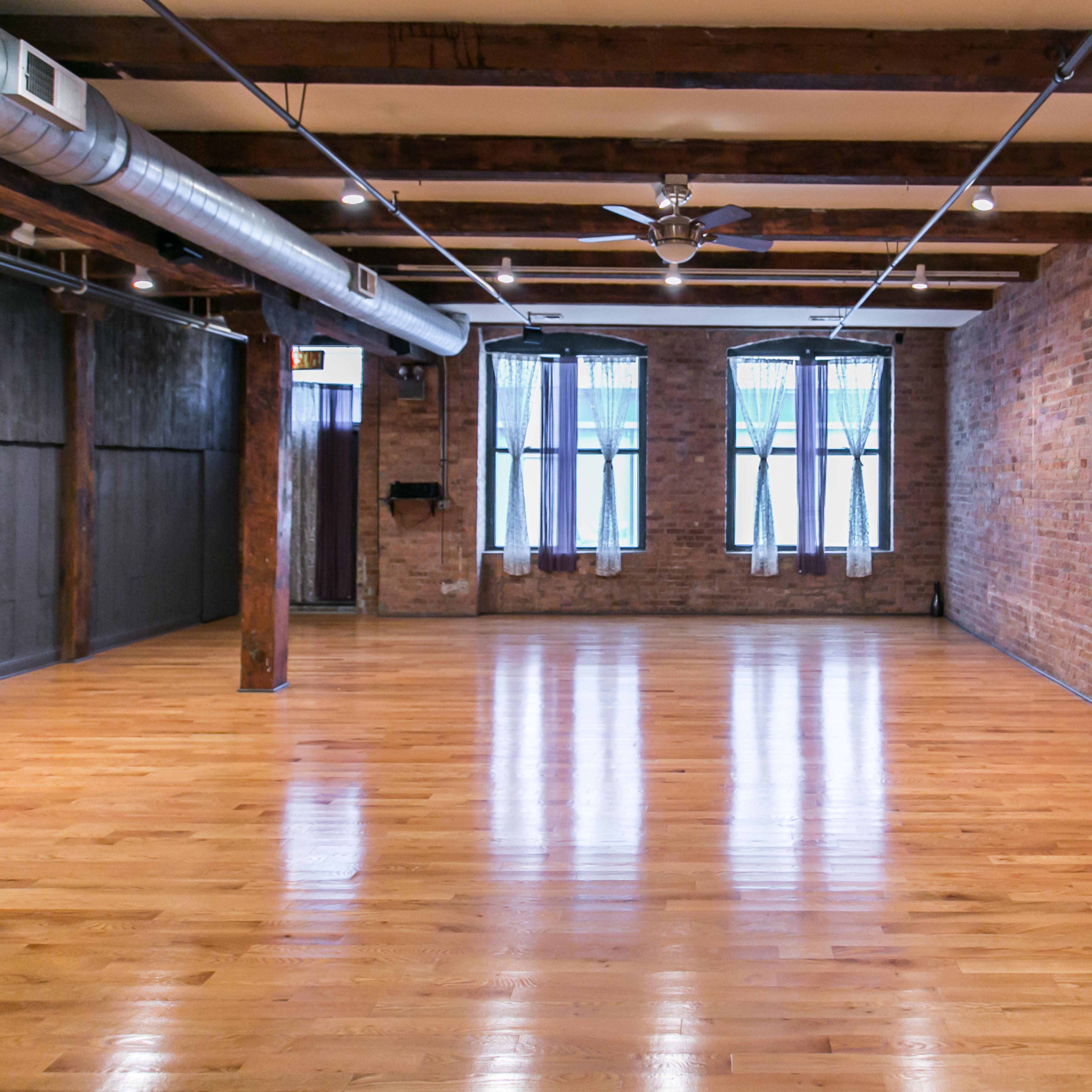 An empty room with exposed brick walls, polished wooden floors, and large windows along one side.