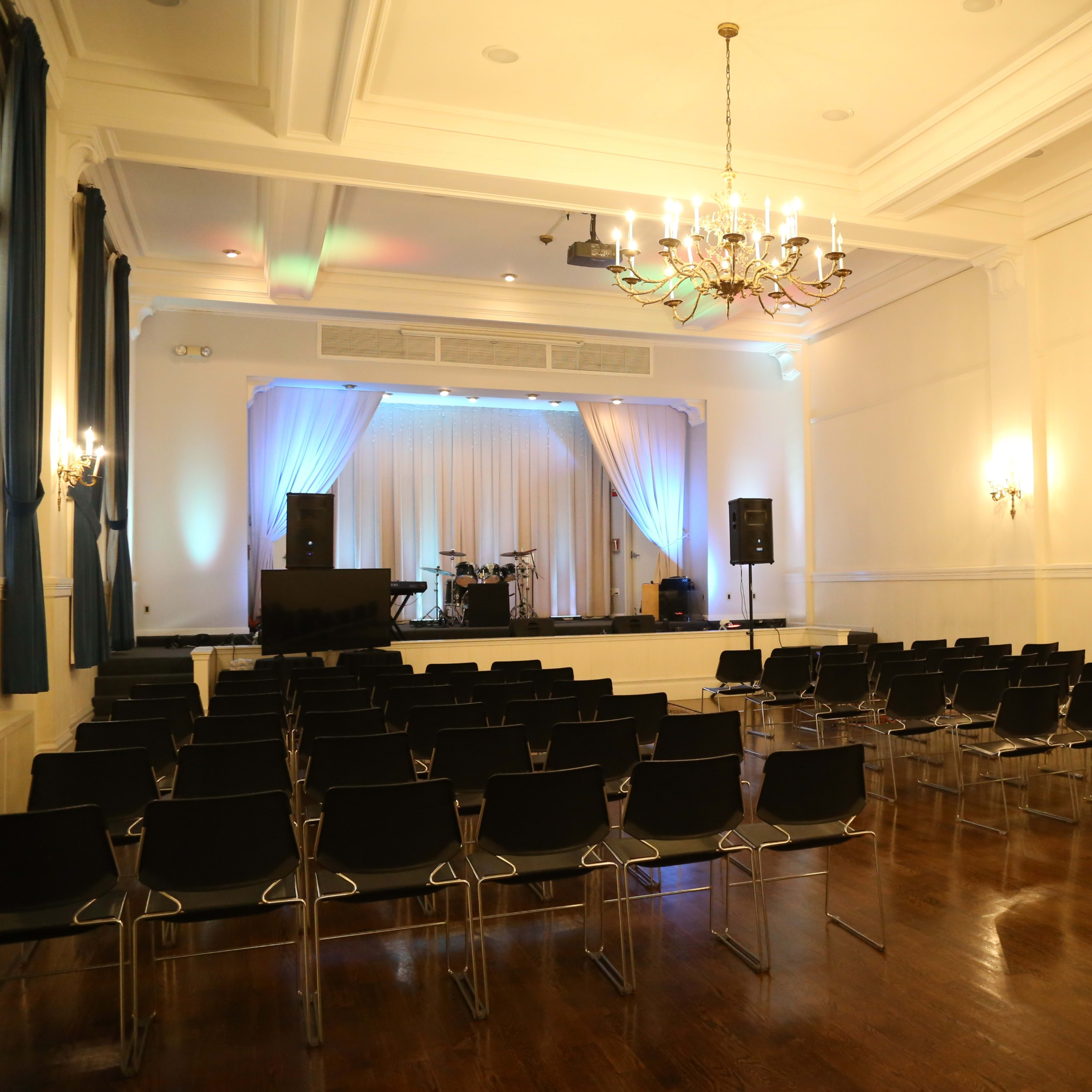 A spacious event hall features rows of black chairs facing a stage with a drum set and sound equipment.