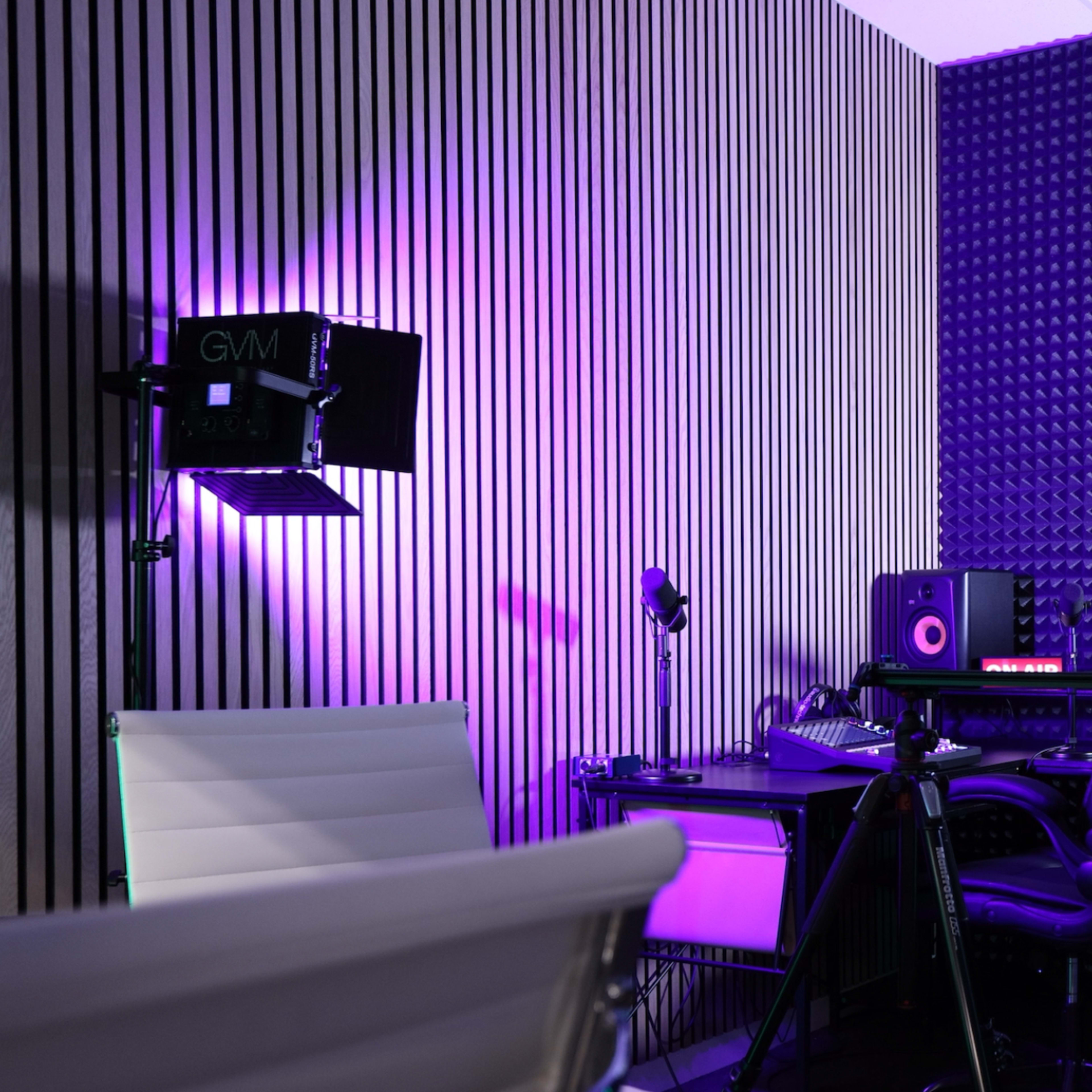 A modern recording studio setup with a desk, ergonomic chair, microphones, monitors, and accent lighting on the walls.