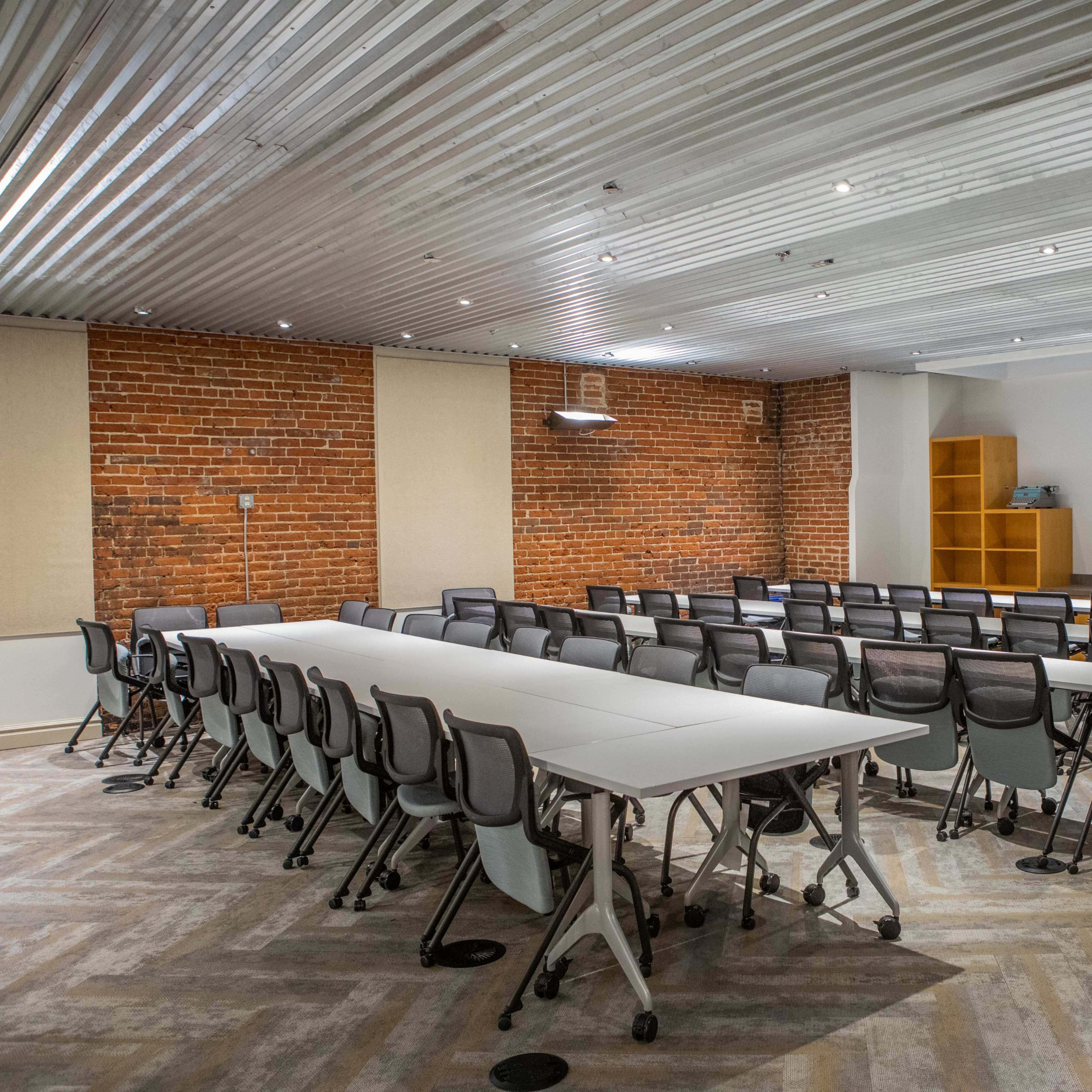 A modern conference room with a long table surrounded by black chairs, exposed brick walls, and a metal ceiling.