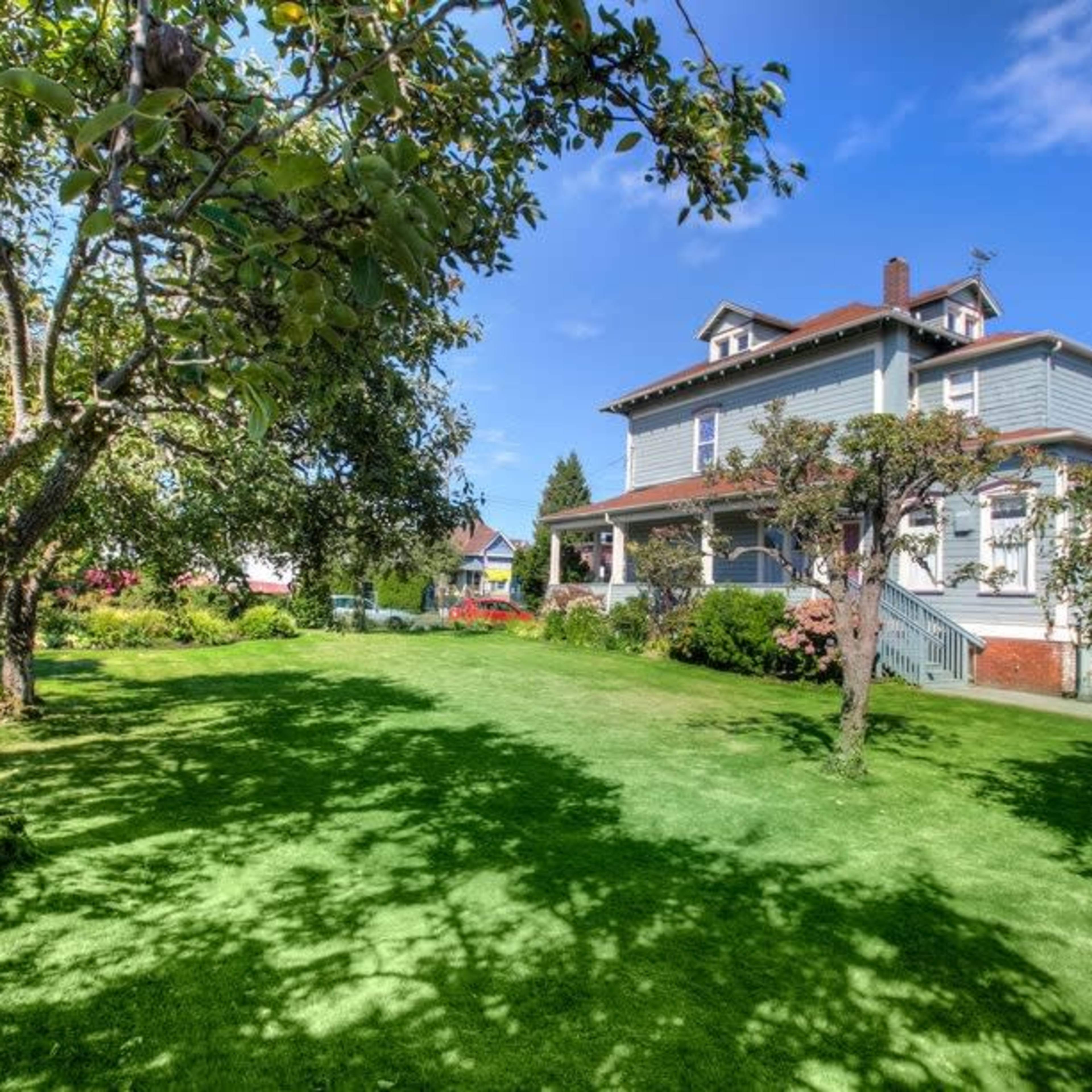 A large house with a Victorian style is surrounded by a grassy yard and several trees under a clear blue sky.