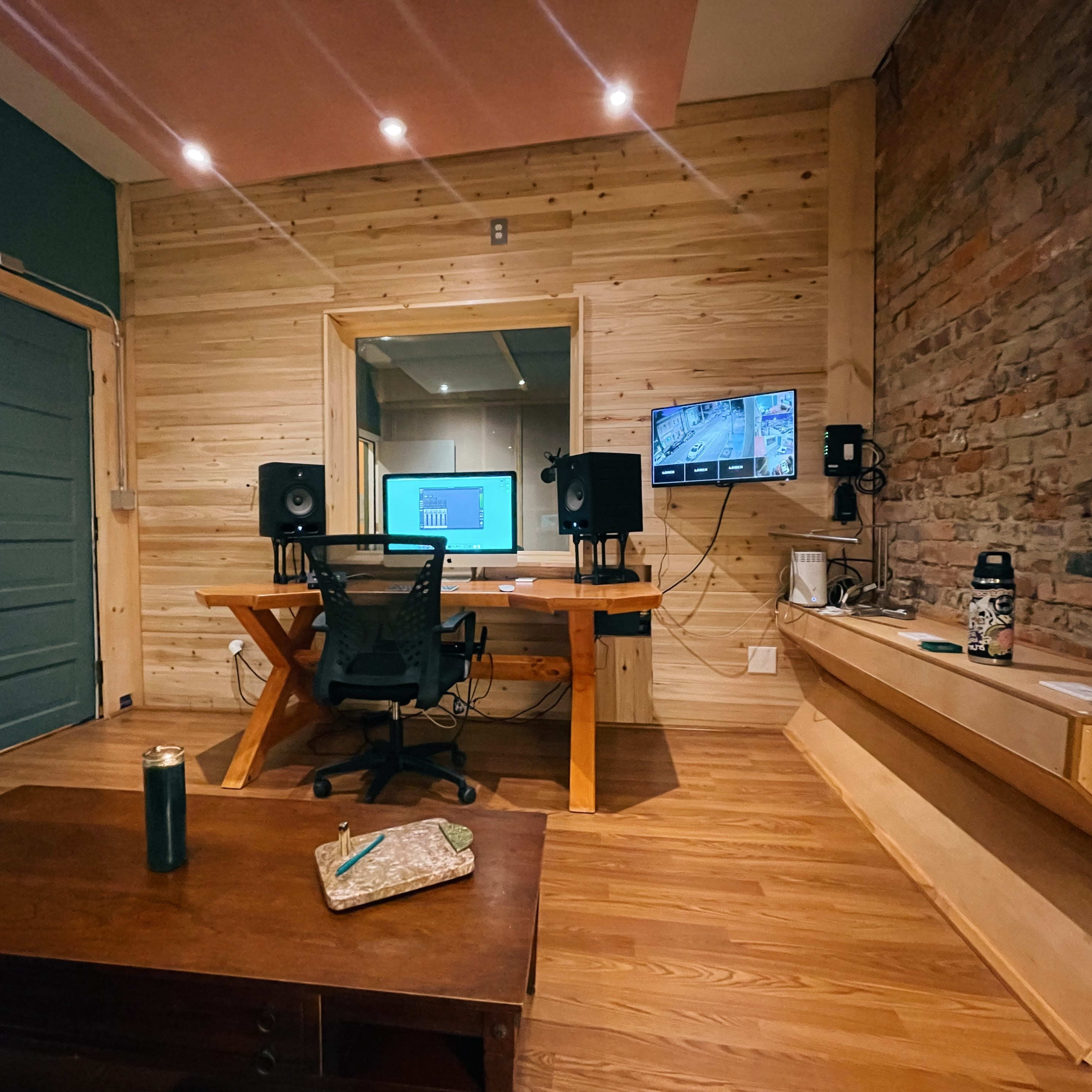 A small recording studio featuring a wooden desk with a computer, audio monitors, and a chair, set against a backdrop of wooden walls and exposed brick.