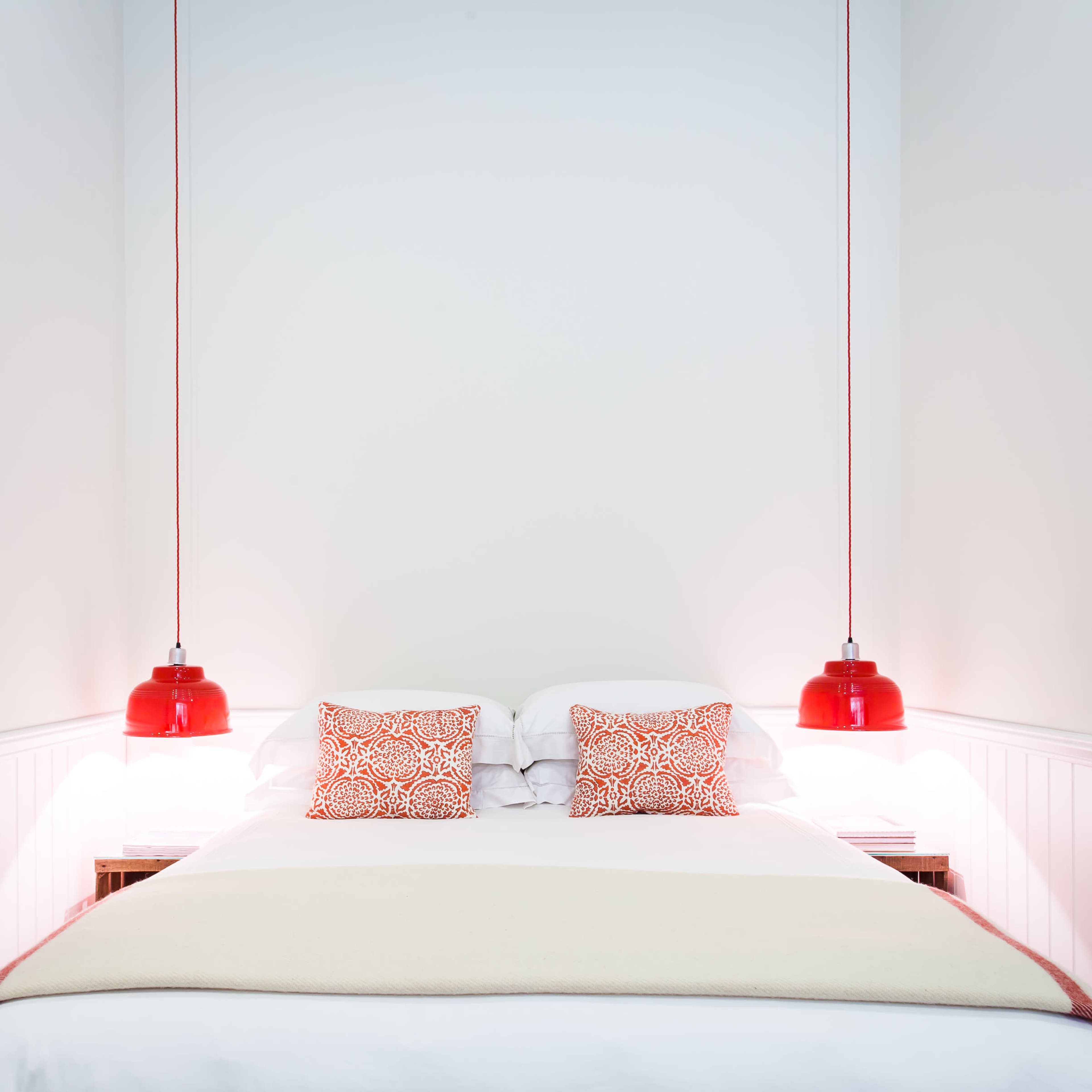 A minimalistic bedroom with a white wall, a bed featuring patterned red and white pillows, and two red pendant lamps on either side.
