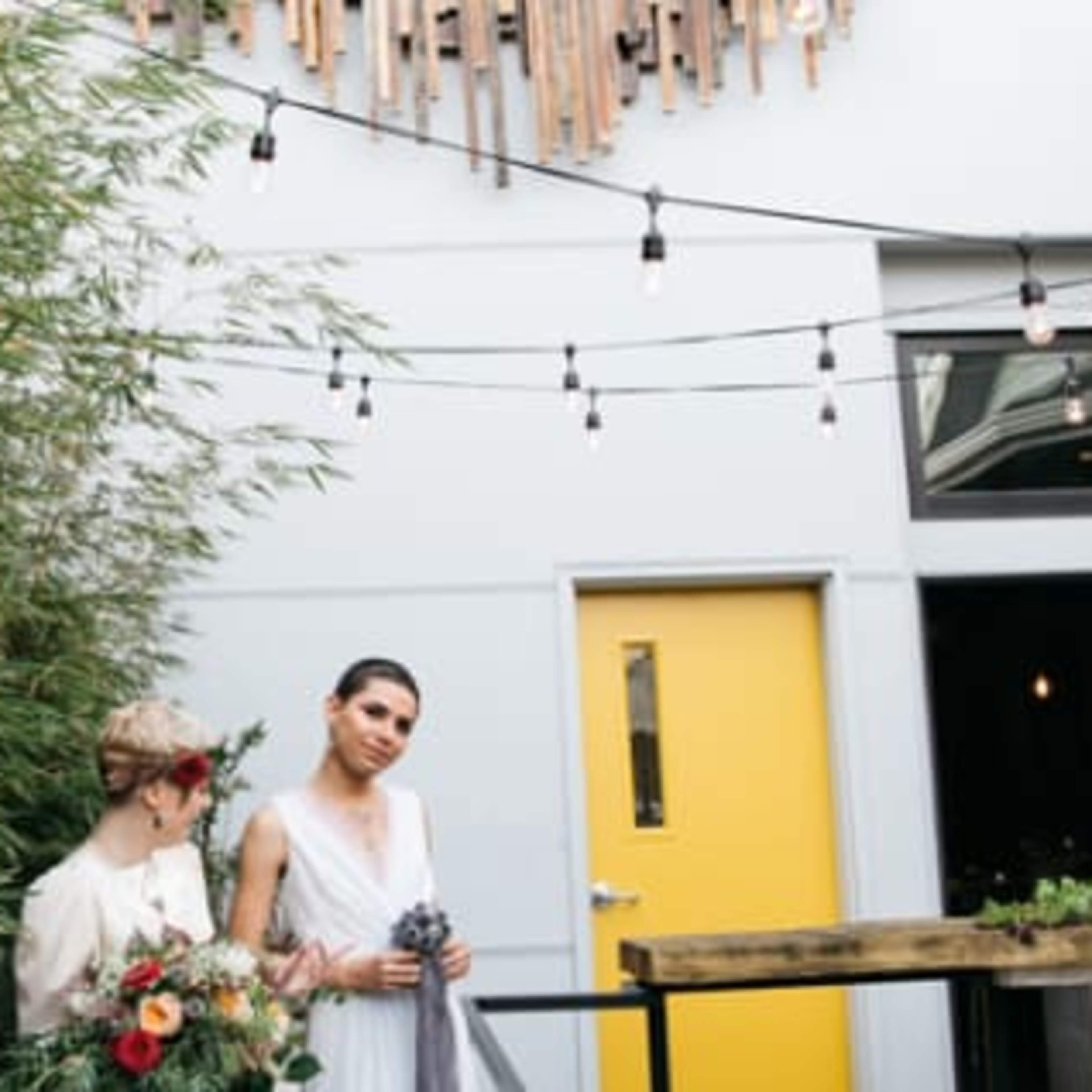 Two women stand in an outdoor space adorned with bamboo and string lights, with one holding a bouquet.
