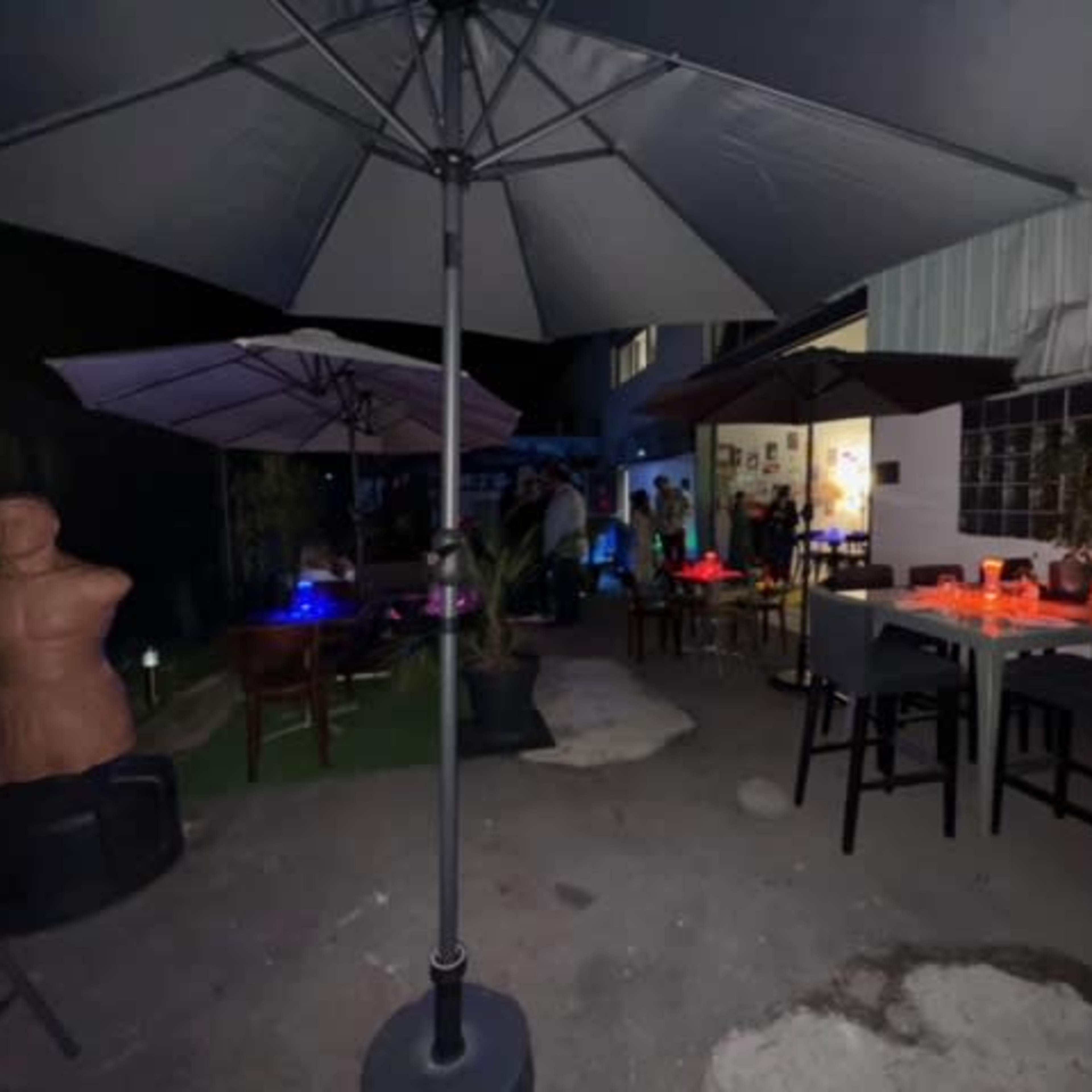 The scene shows a nighttime outdoor area with tables, umbrellas, and a dummy figure, set up for a gathering.