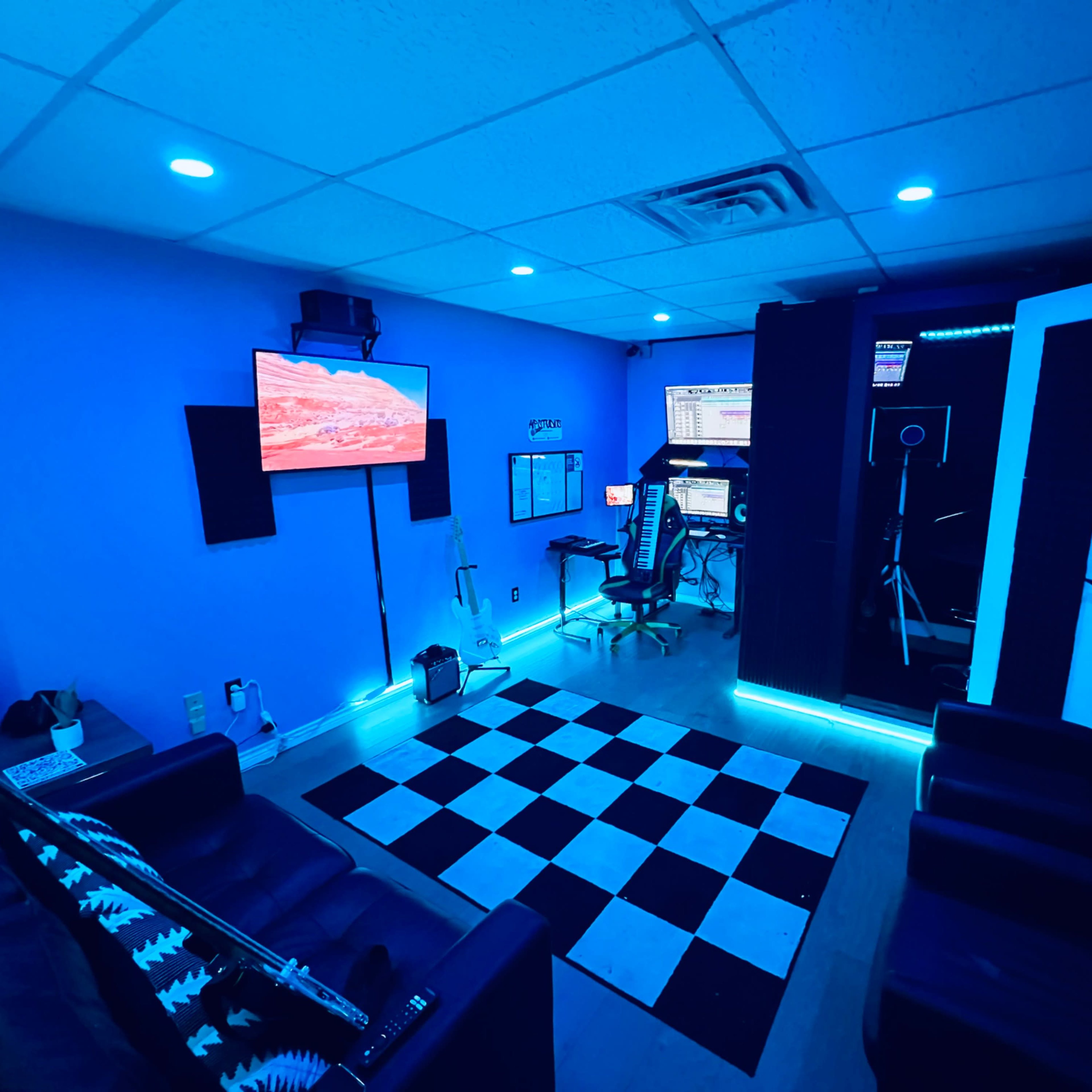 A room with a checkered floor, two black couches, a wall-mounted TV, and computer equipment against blue-lit walls.