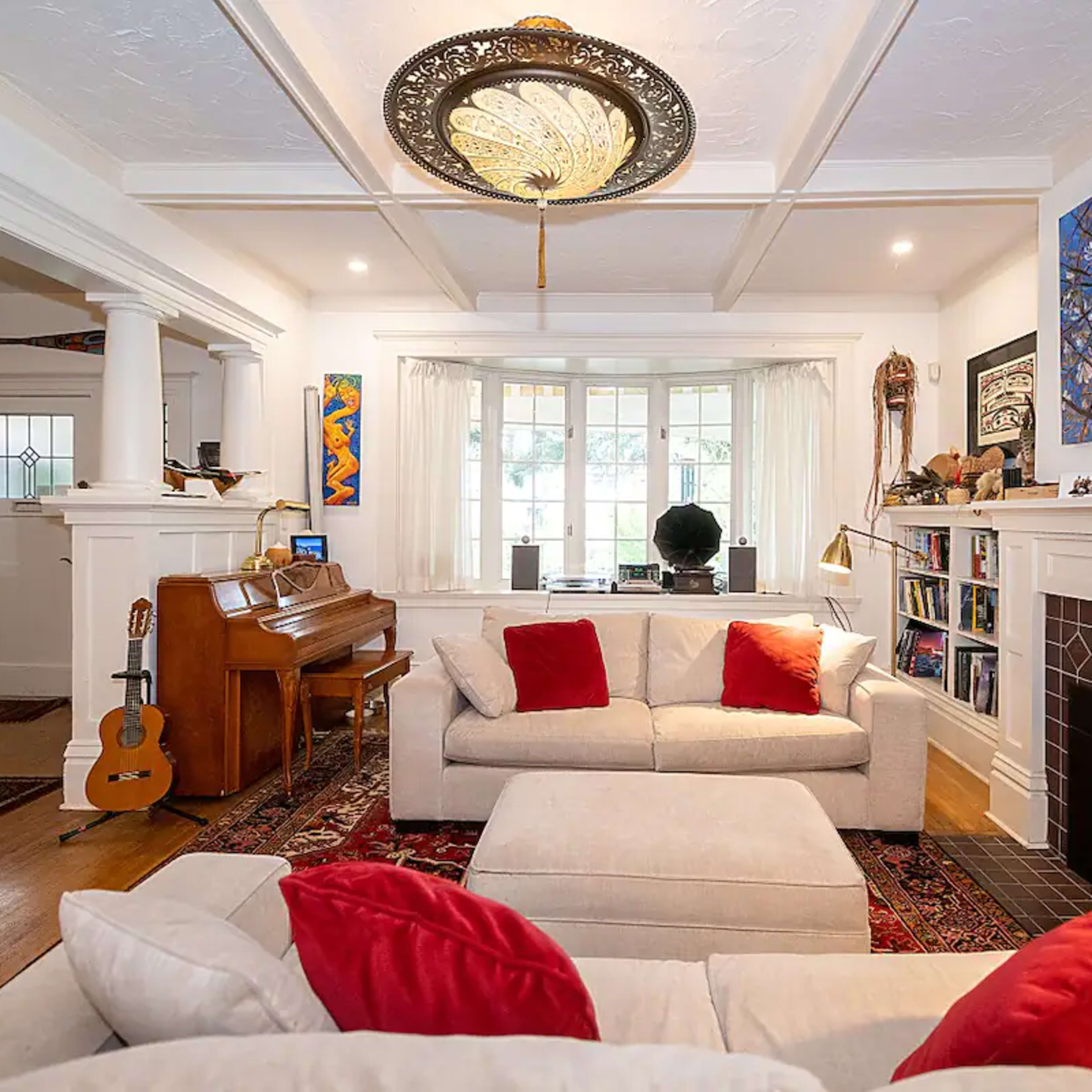 The living room features a piano, two light-colored sofas with red pillows, a fireplace, and colorful artwork on the walls.