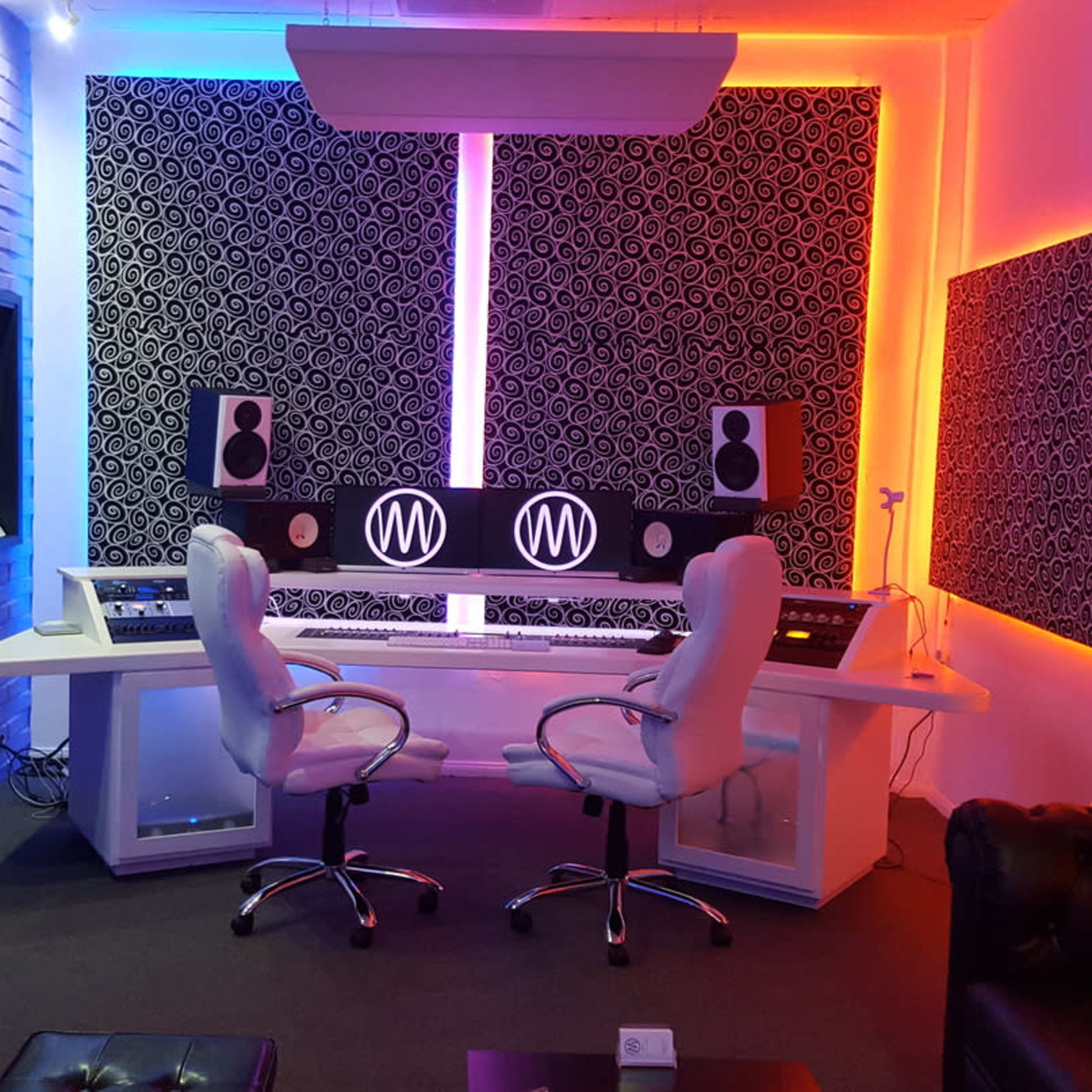 A modern music studio featuring two white chairs at a double workstation, surrounded by colorful LED lighting and soundproof wall panels.