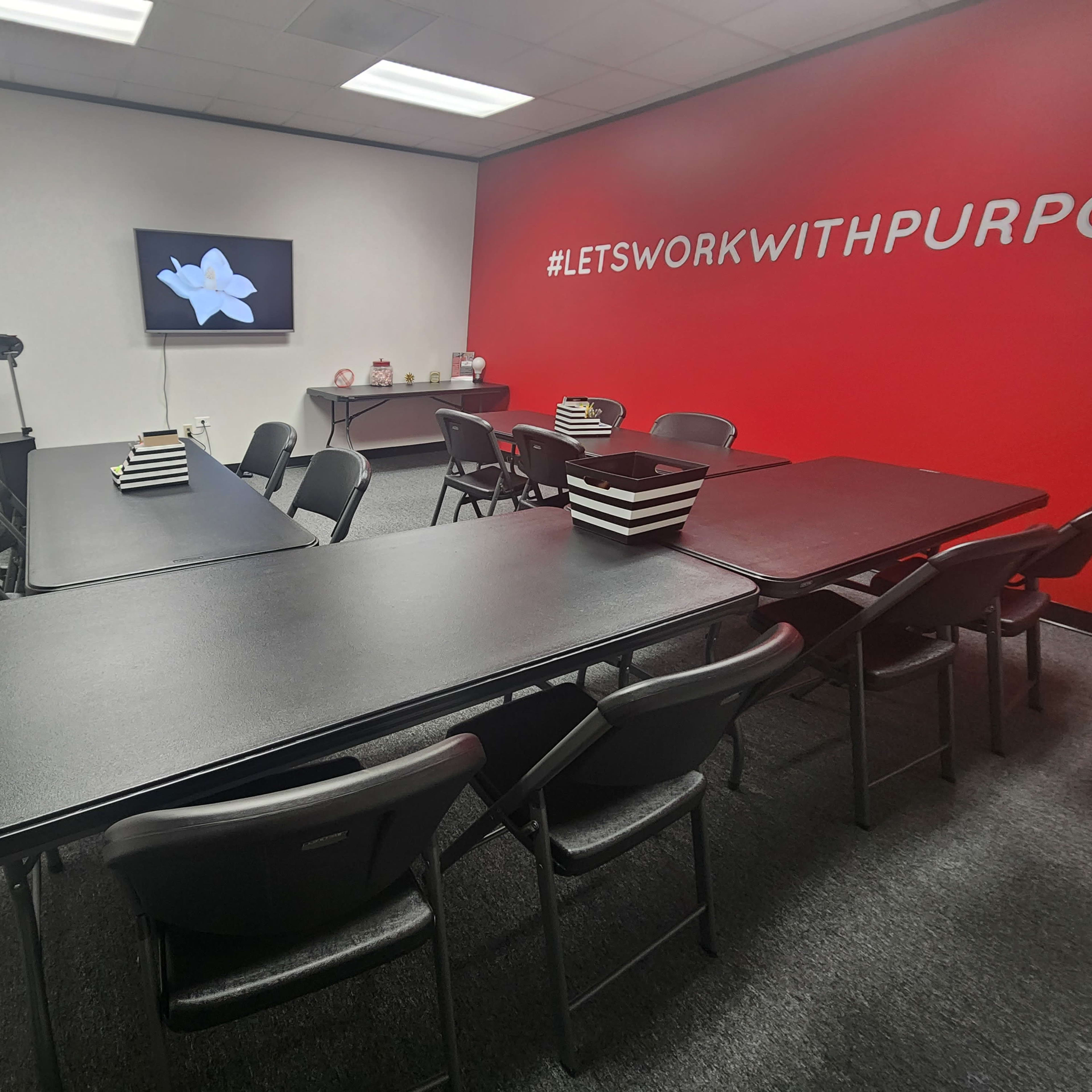 A conference room is set up with several black tables and chairs, featuring a wall-mounted screen displaying a flower image.
