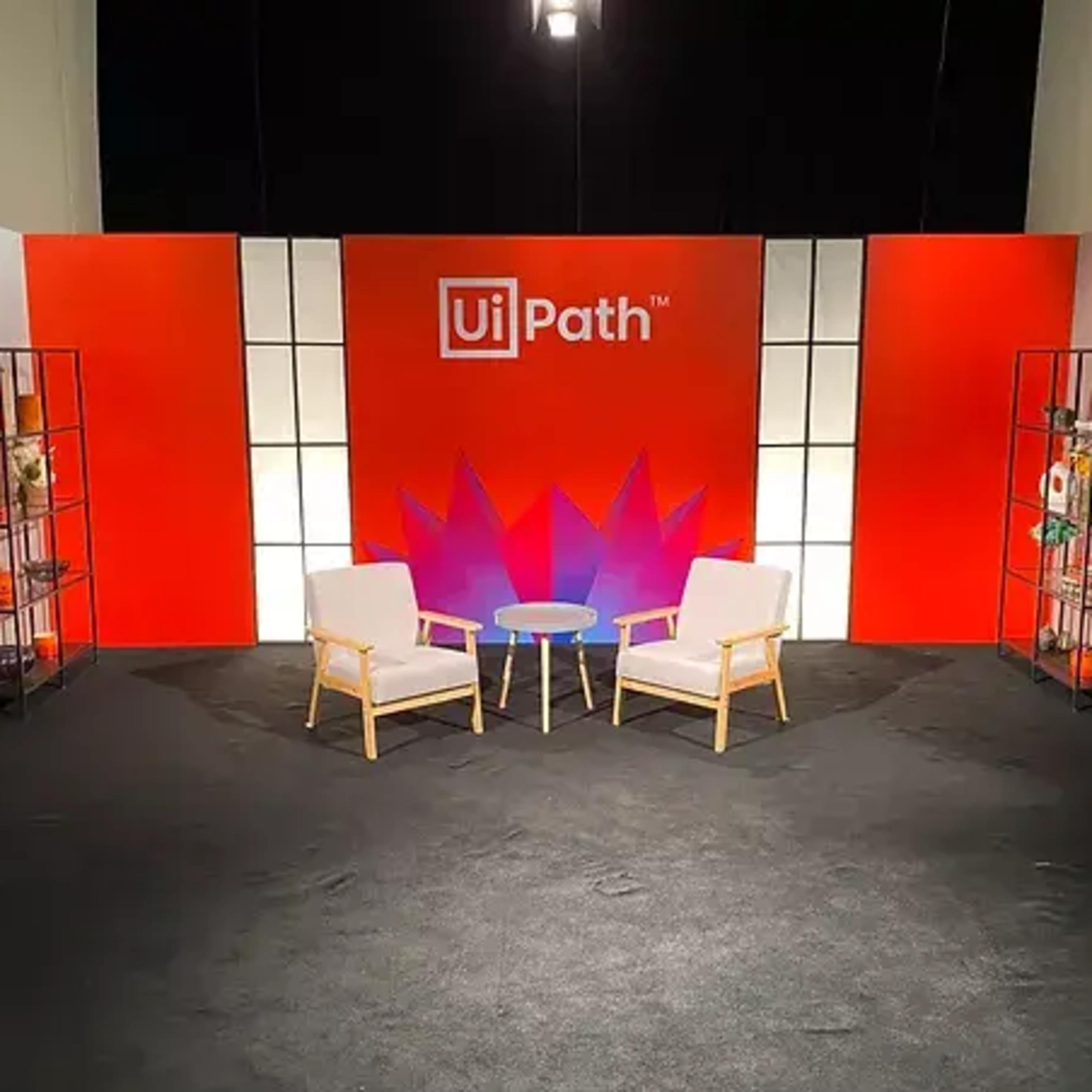 A stage setup with two white chairs and a round table in front of a vibrant backdrop featuring the UiPath logo and decorative elements.