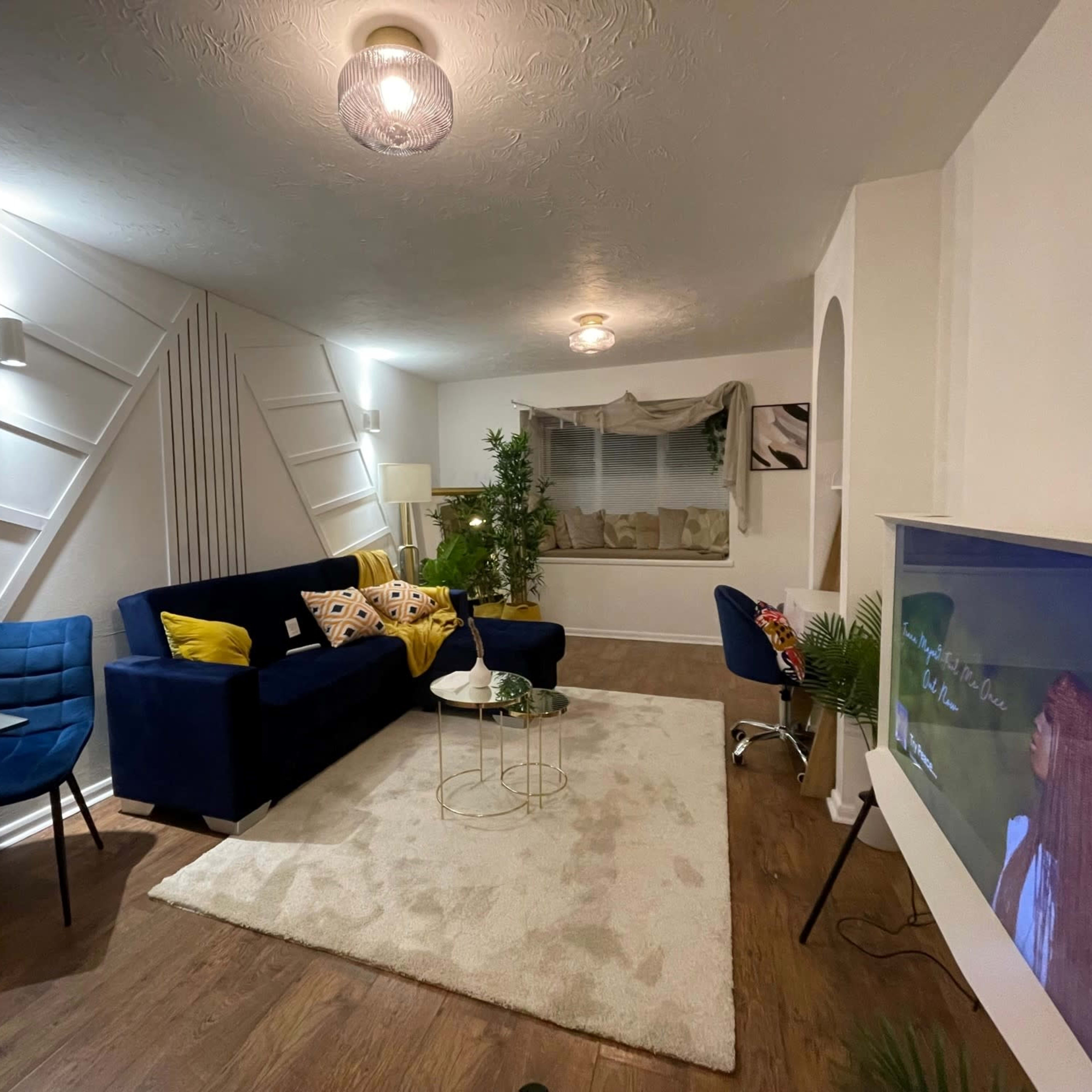 A modern living room featuring a blue sofa, a glass table, and a television mounted on the wall, with a decorative rug on the floor and plants for accents.