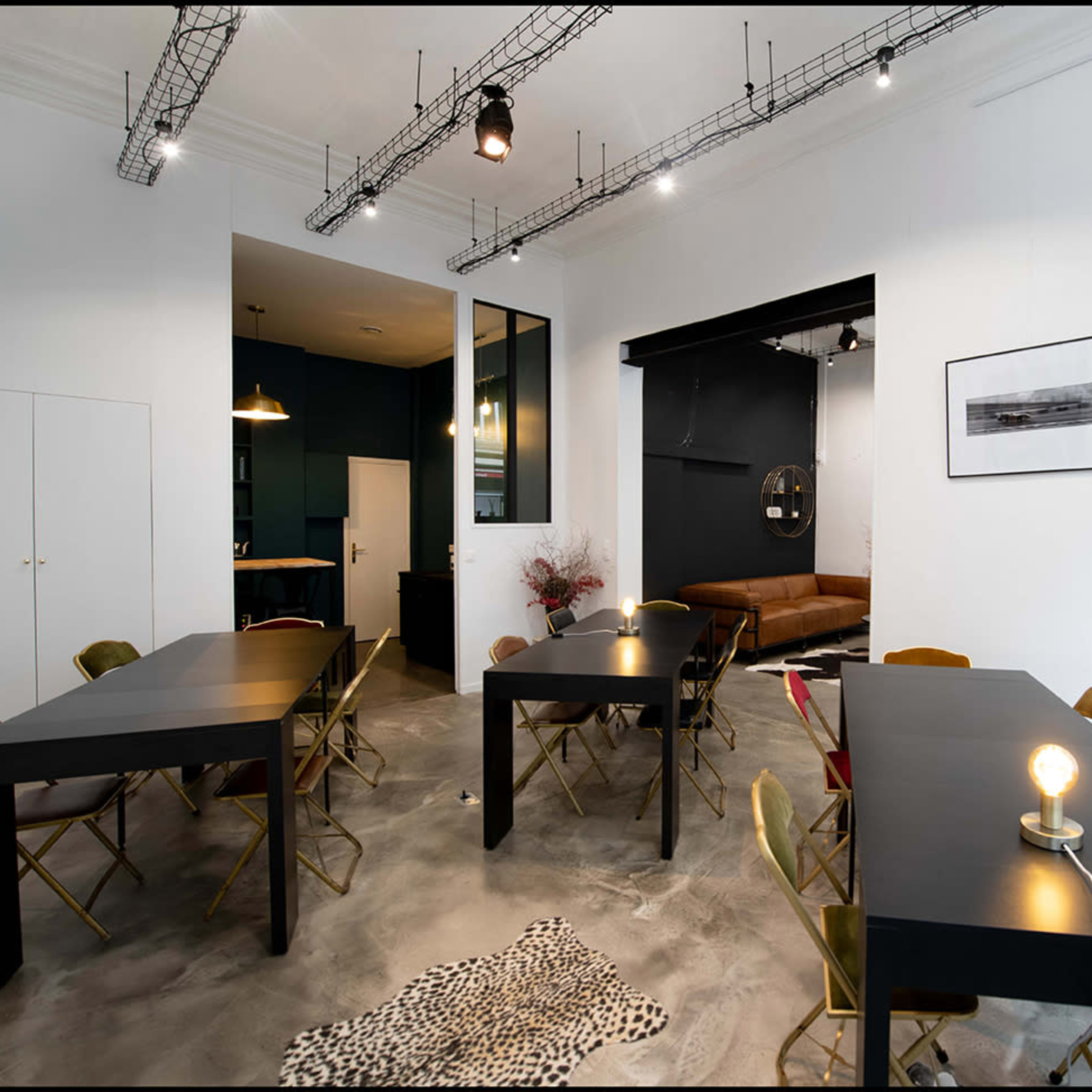 A modern workspace featuring several black tables with golden chairs, a sofa in the background, and minimalist decor on the walls.