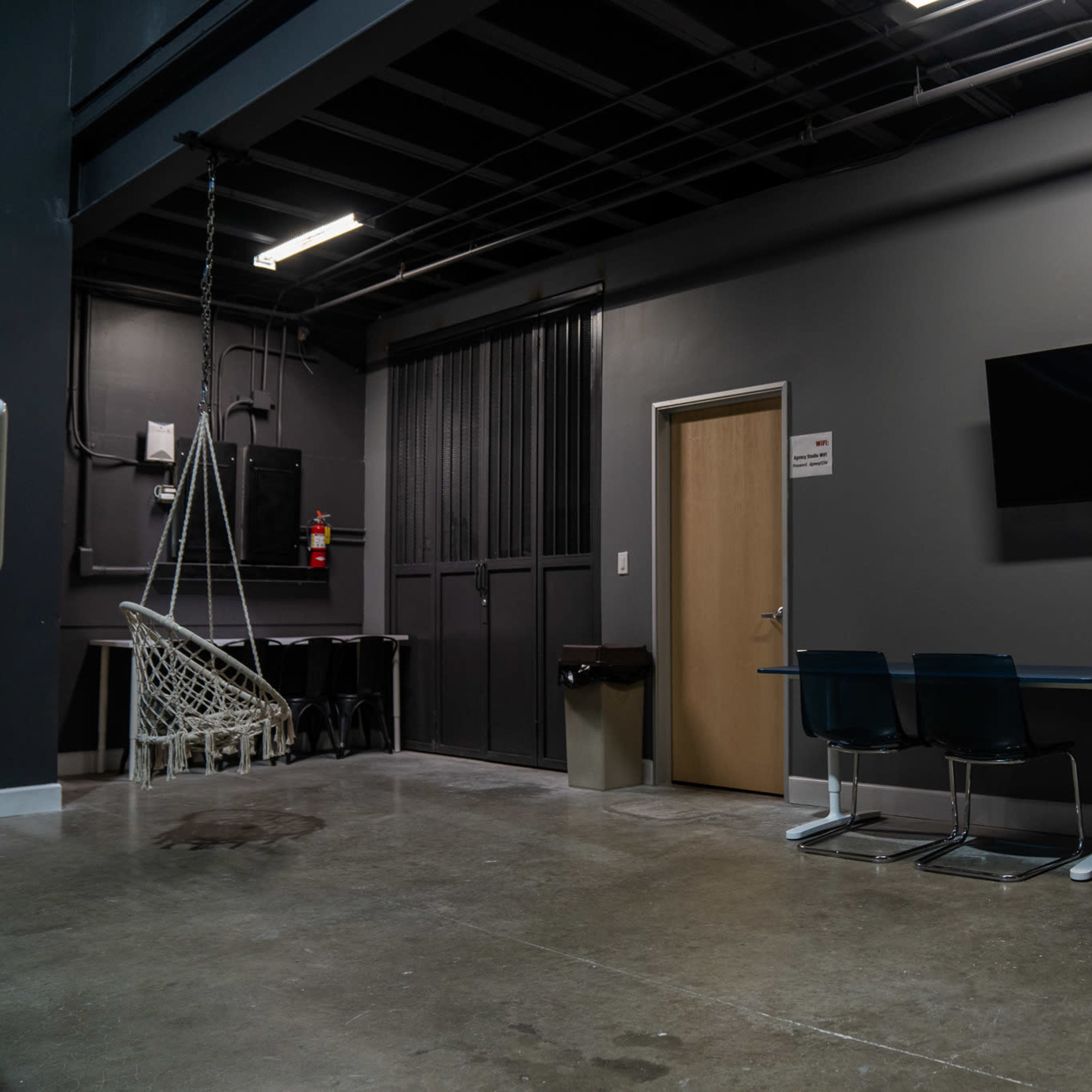 A minimalist room with gray walls, a few chairs, a table, a television mounted on the wall, and a rope swing in one corner.
