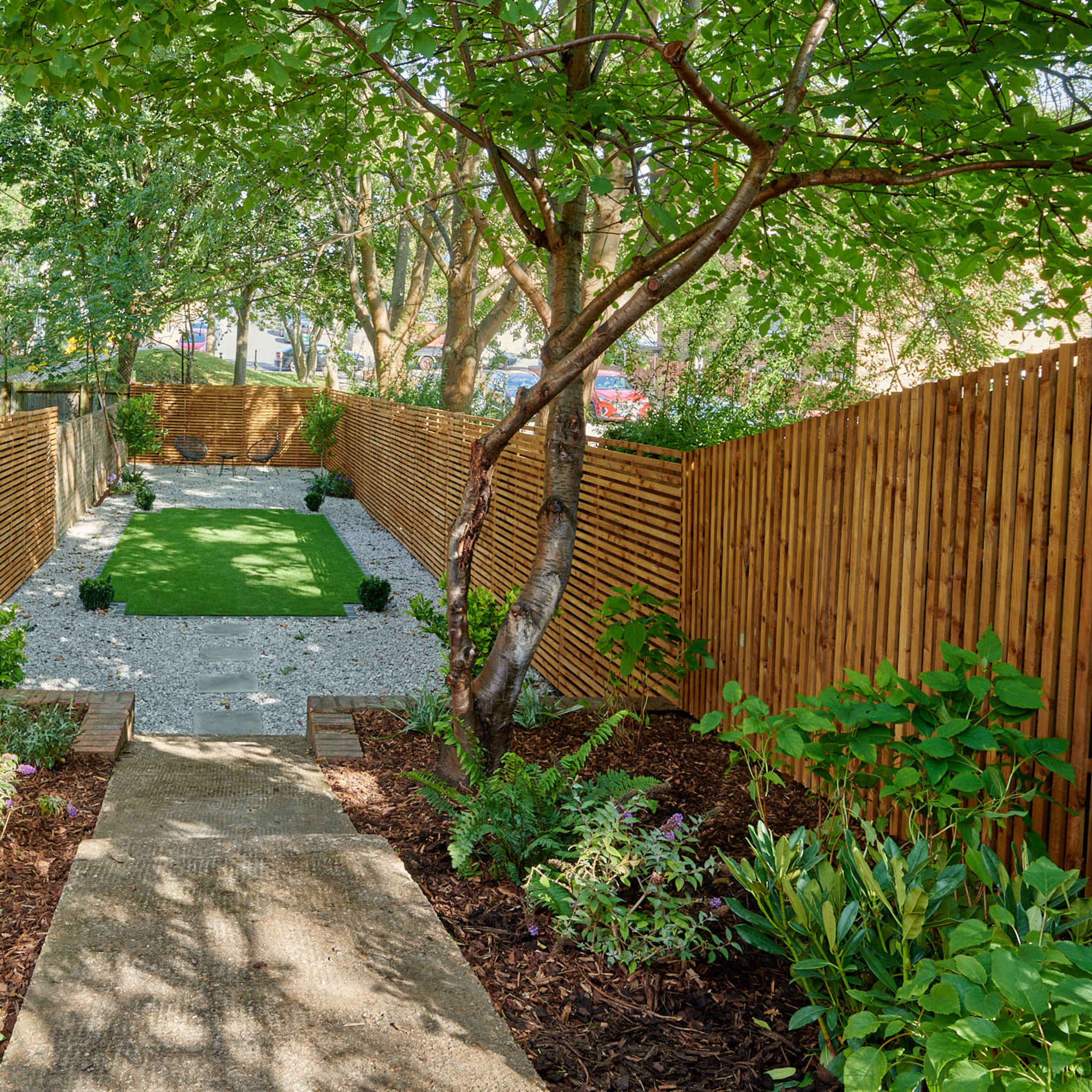 A narrow garden pathway leading to a green lawn bordered by wooden fencing and landscaped with various plants and gravel.