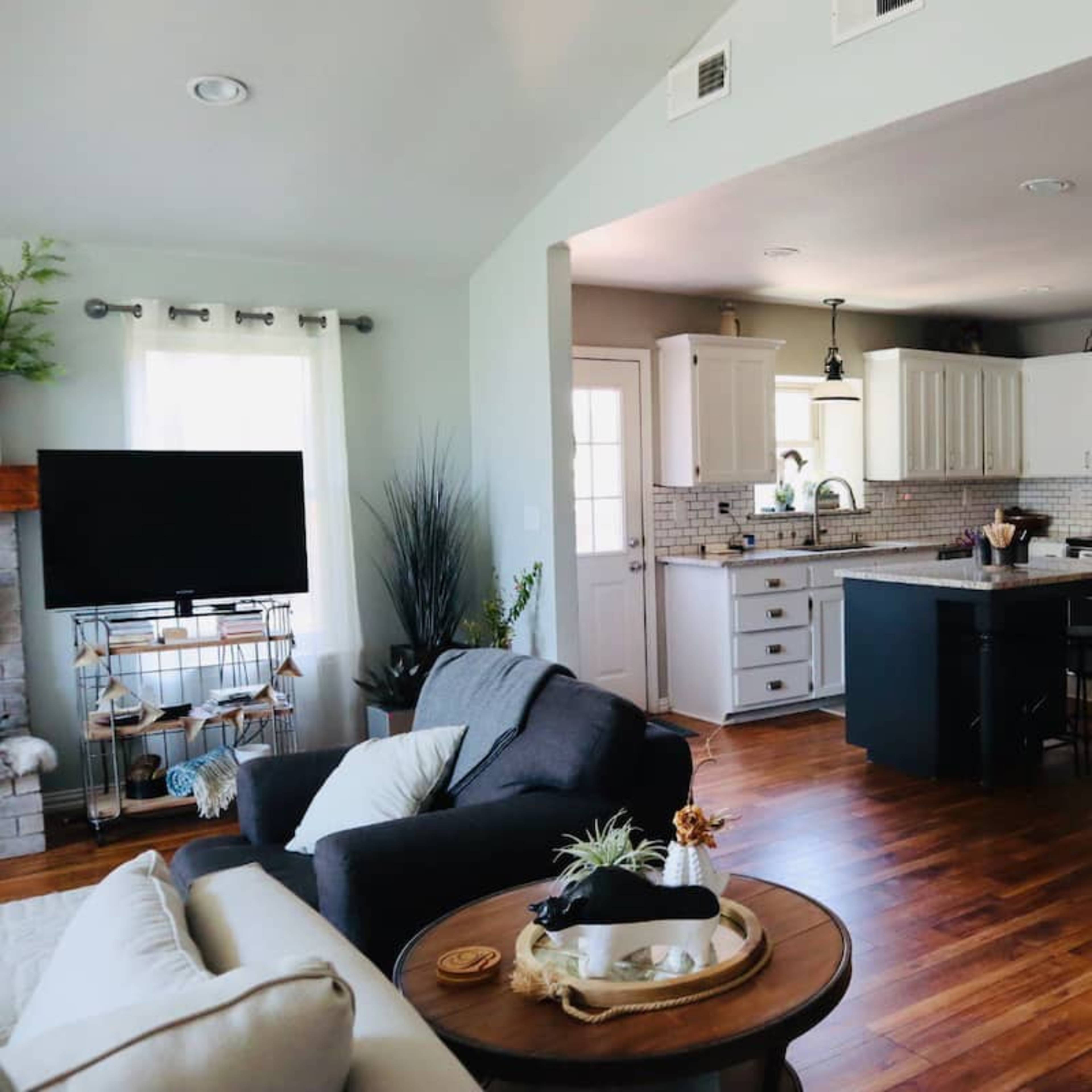 A modern kitchen and living area with wooden flooring, white cabinetry, a gray island, and a dining table set for meals.