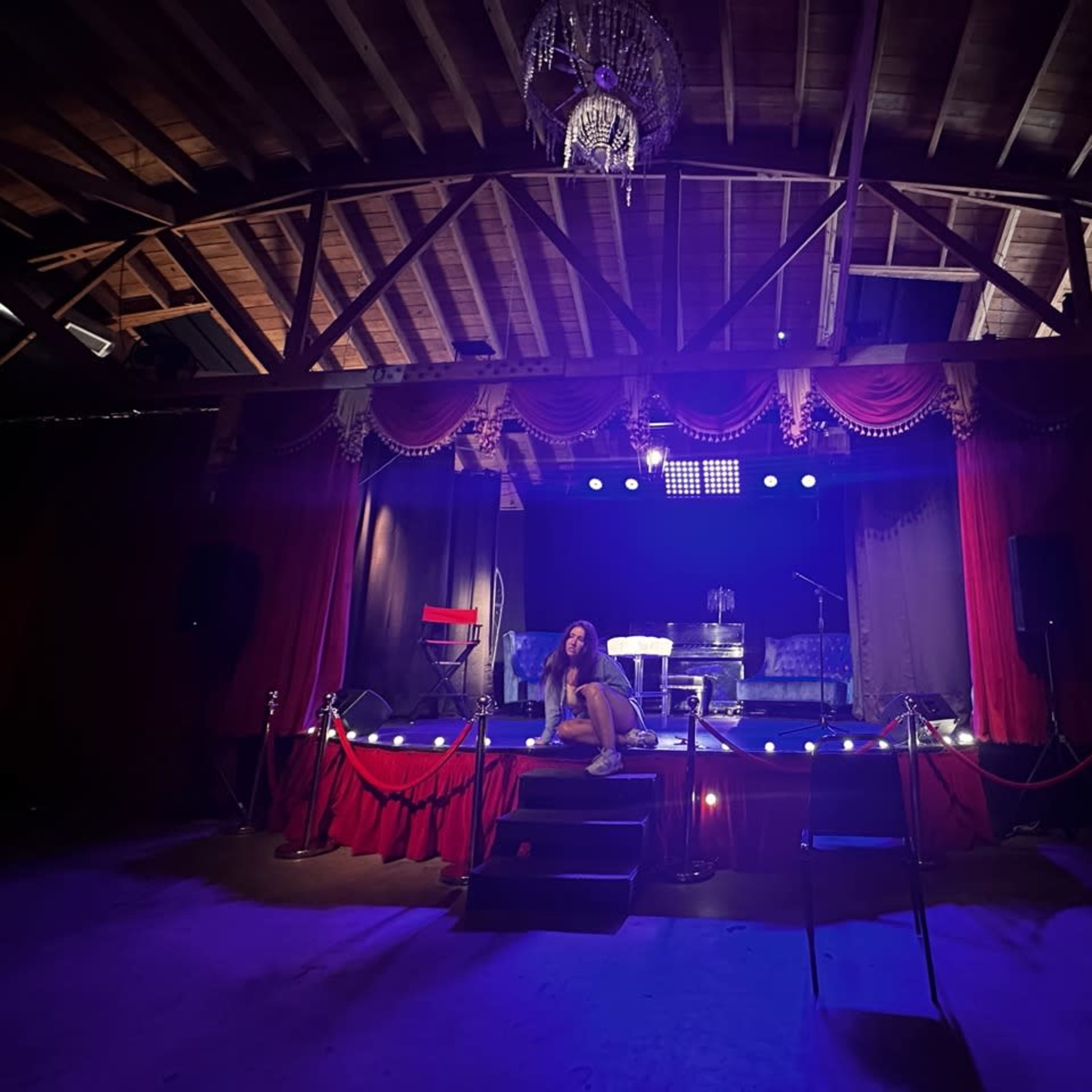 A performer sits on the steps of a dimly lit stage decorated with red curtains, a chandelier overhead, and various spotlights illuminating the scene.