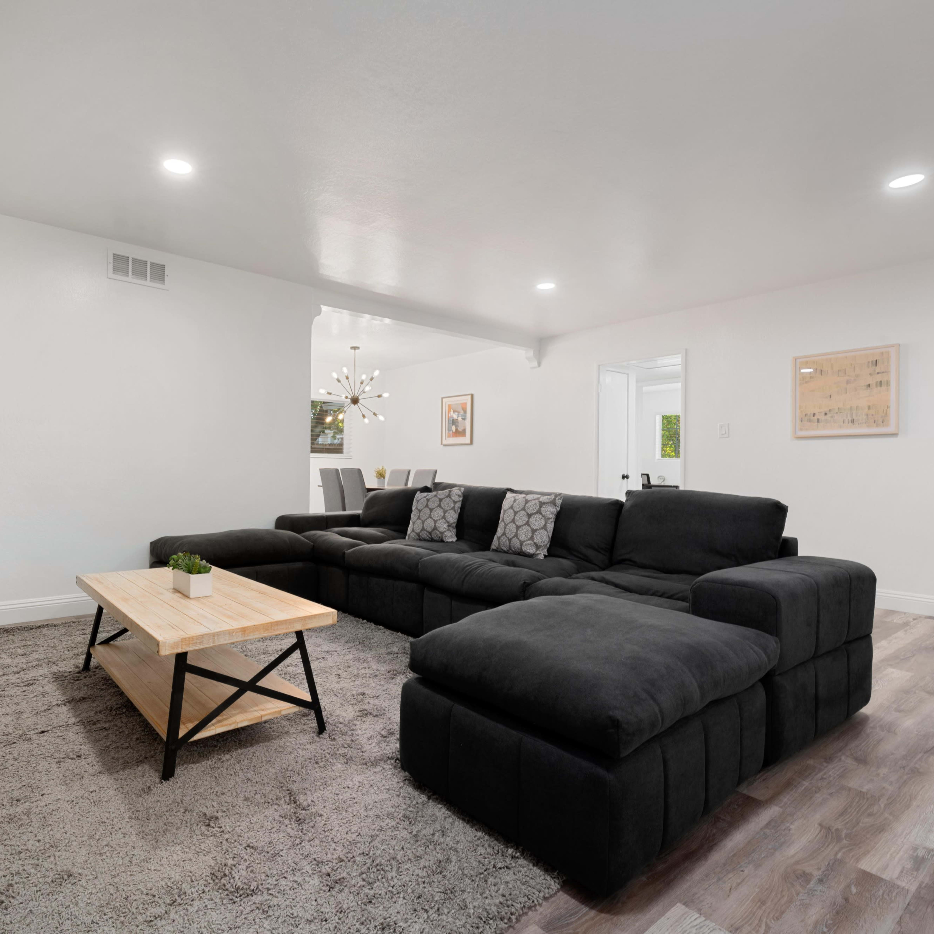A modern living room with a large black sectional sofa, a wooden coffee table, and a plush area rug, illuminated by ceiling lights.