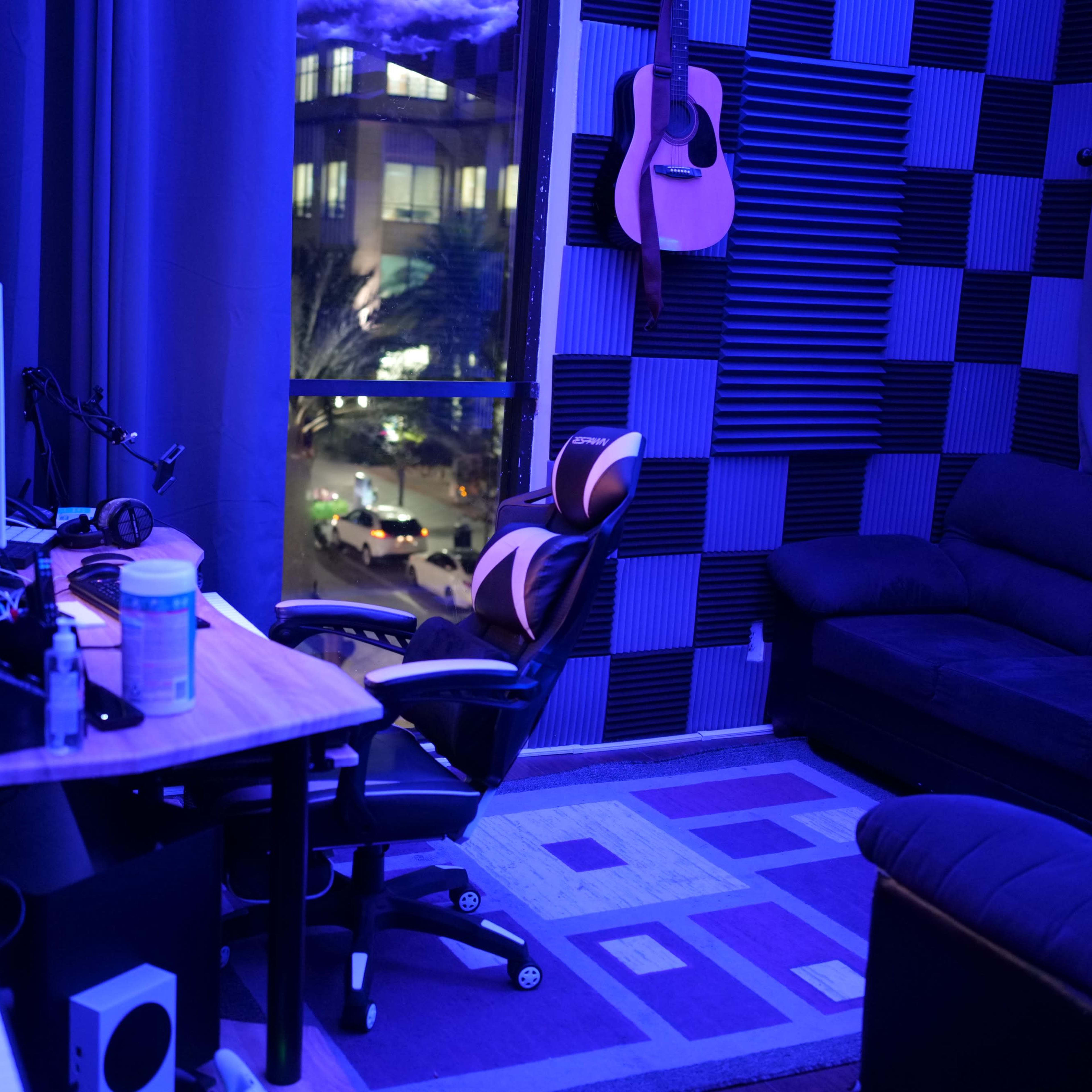 A dimly lit music studio with blue-tinted walls, a desk with computer equipment, a gaming chair, and a sofa, all adjacent to a window revealing a cityscape at night.