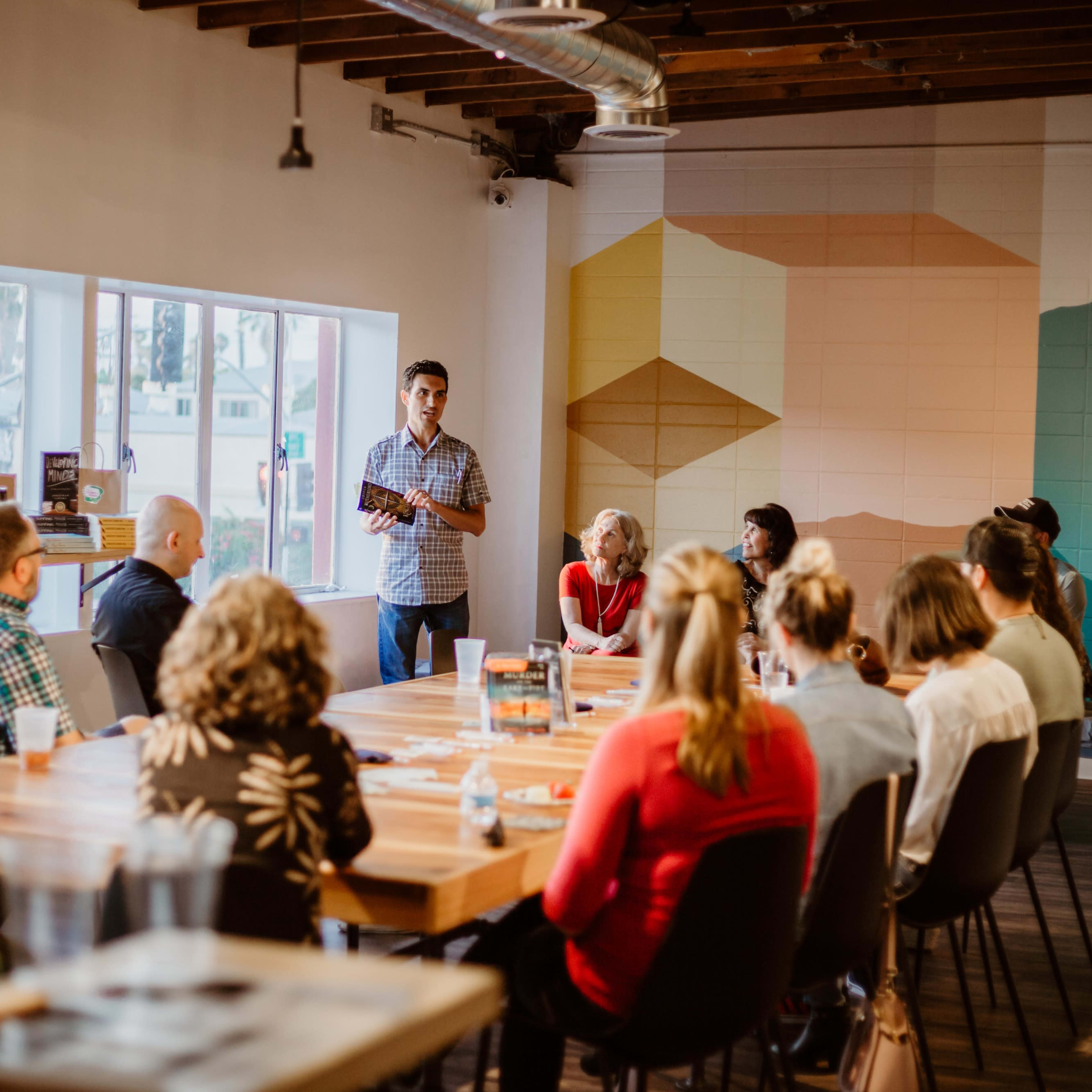 A speaker stands in front of a group seated around a large wooden table in a well-lit meeting space.