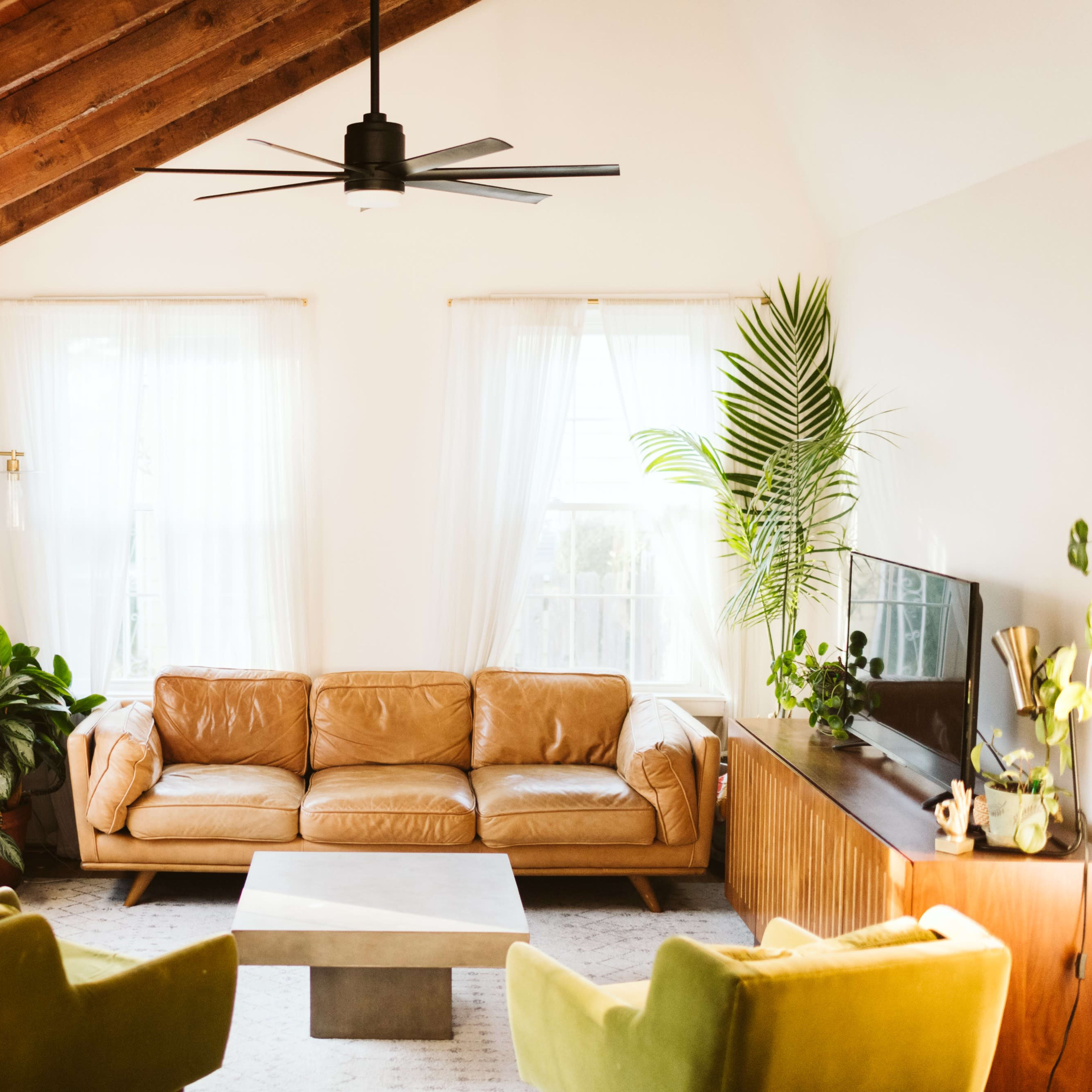 A modern living room features a brown leather sofa, two green armchairs, a coffee table, and large windows with sheer curtains.