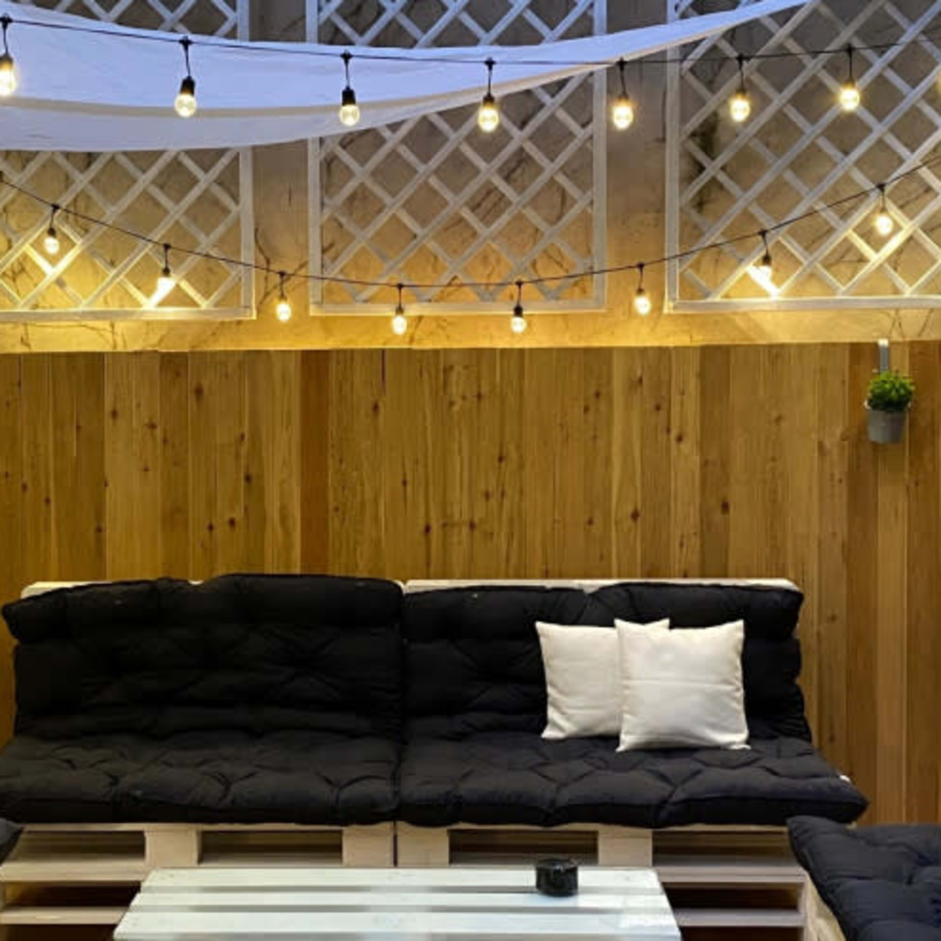 A cozy outdoor seating area with dark cushioned sofas arranged around a white coffee table, illuminated by string lights and draped fabric overhead.