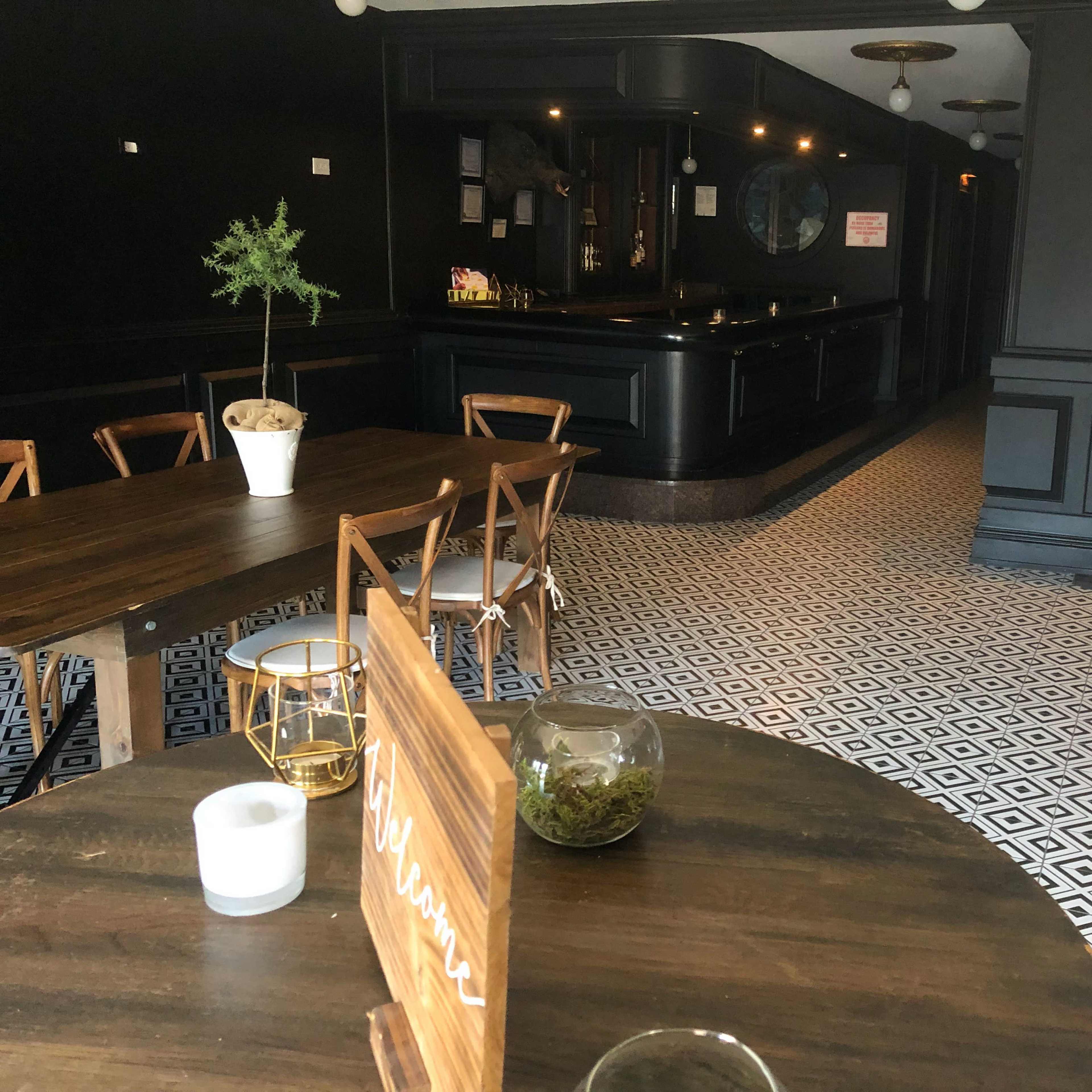 A stylish interior of a café or restaurant featuring wooden tables, a black bar area, and geometric-patterned flooring.