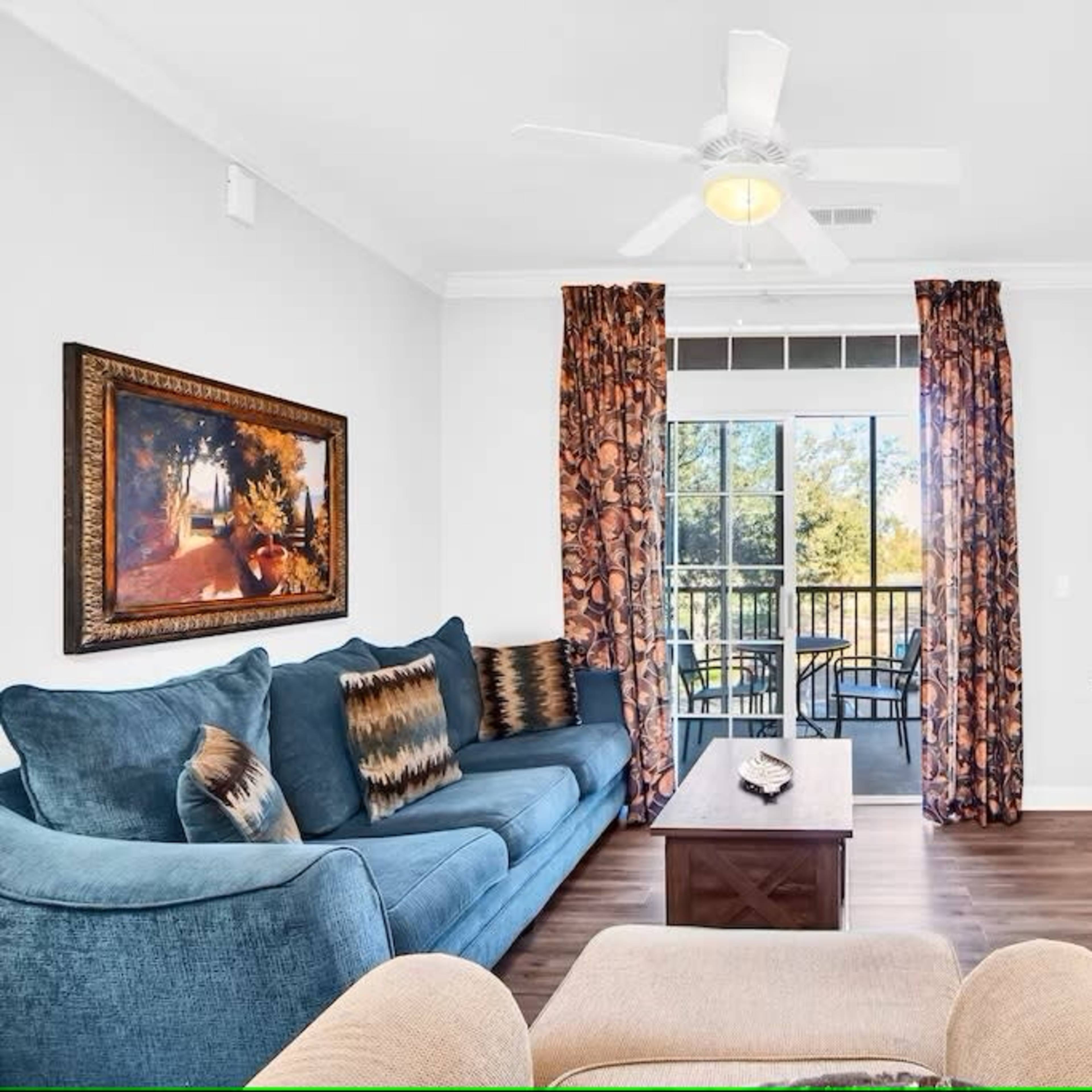 A living room features a blue sofa, a wooden coffee table, a television mounted on the wall, and curtains with floral patterns, overlooking a balcony through large windows.