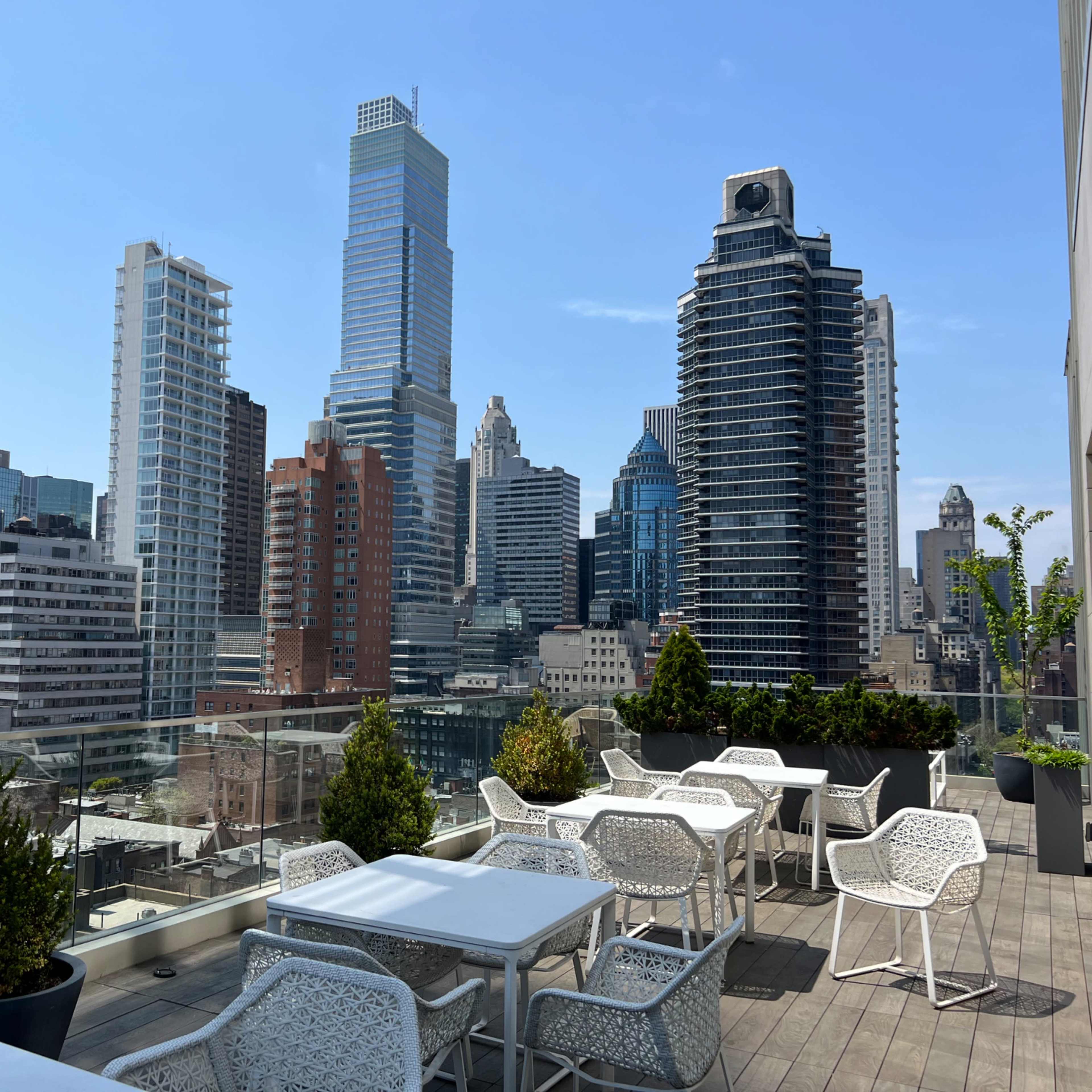 A rooftop terrace with white tables and chairs, overlooking a skyline of tall buildings in a city.