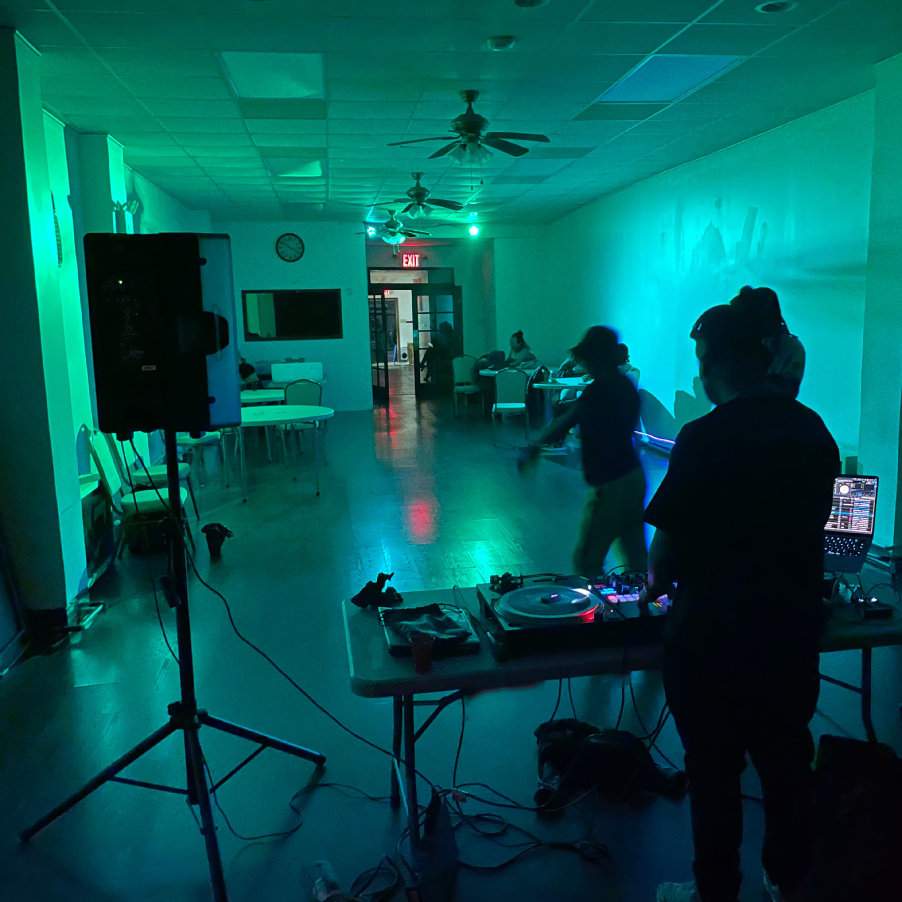 A DJ stands behind a table with turntables and a laptop as a person dances in a dimly lit room with green lights.