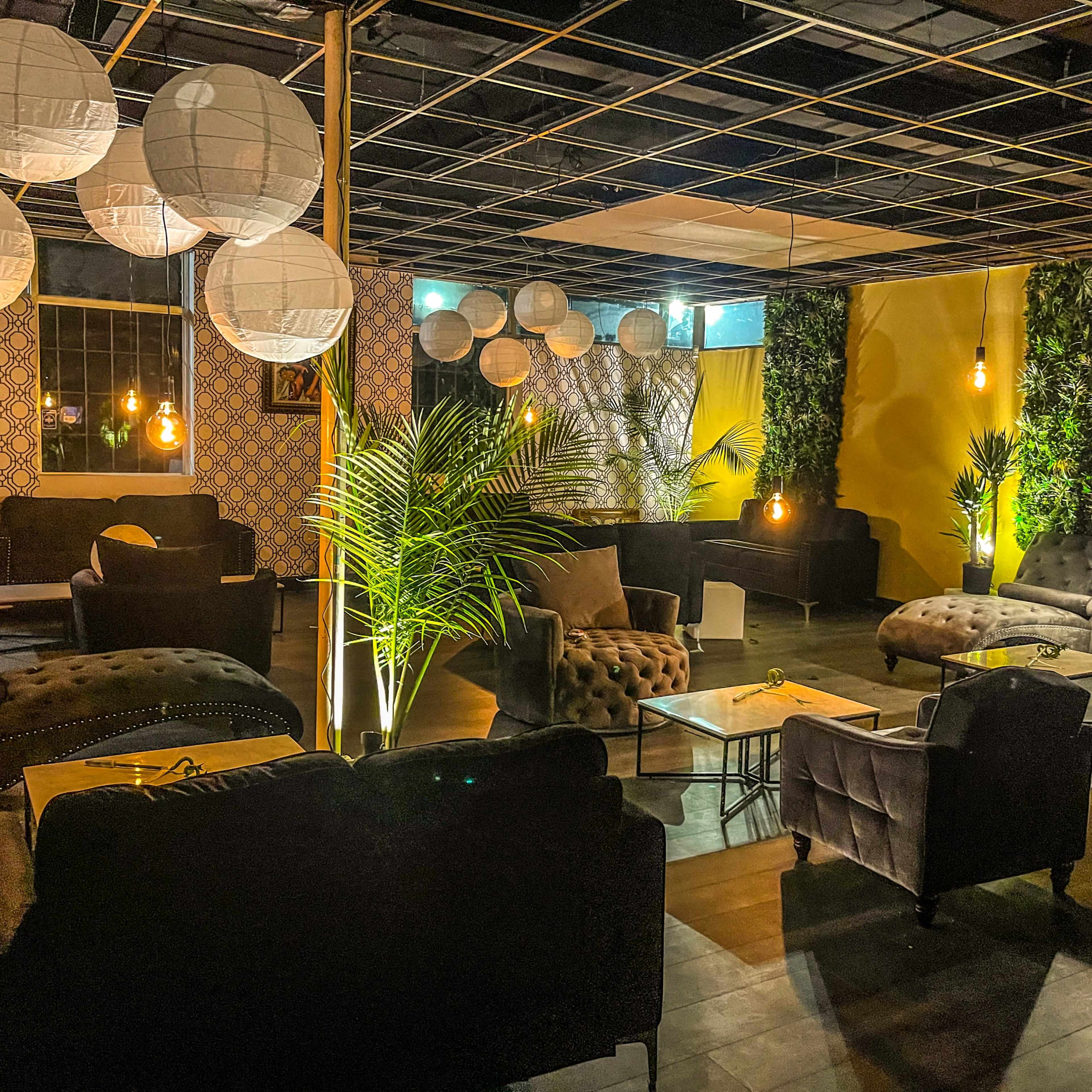 A modern lounge area featuring dark furniture, decorative plants, and soft lighting from pendant lamps and paper lanterns.