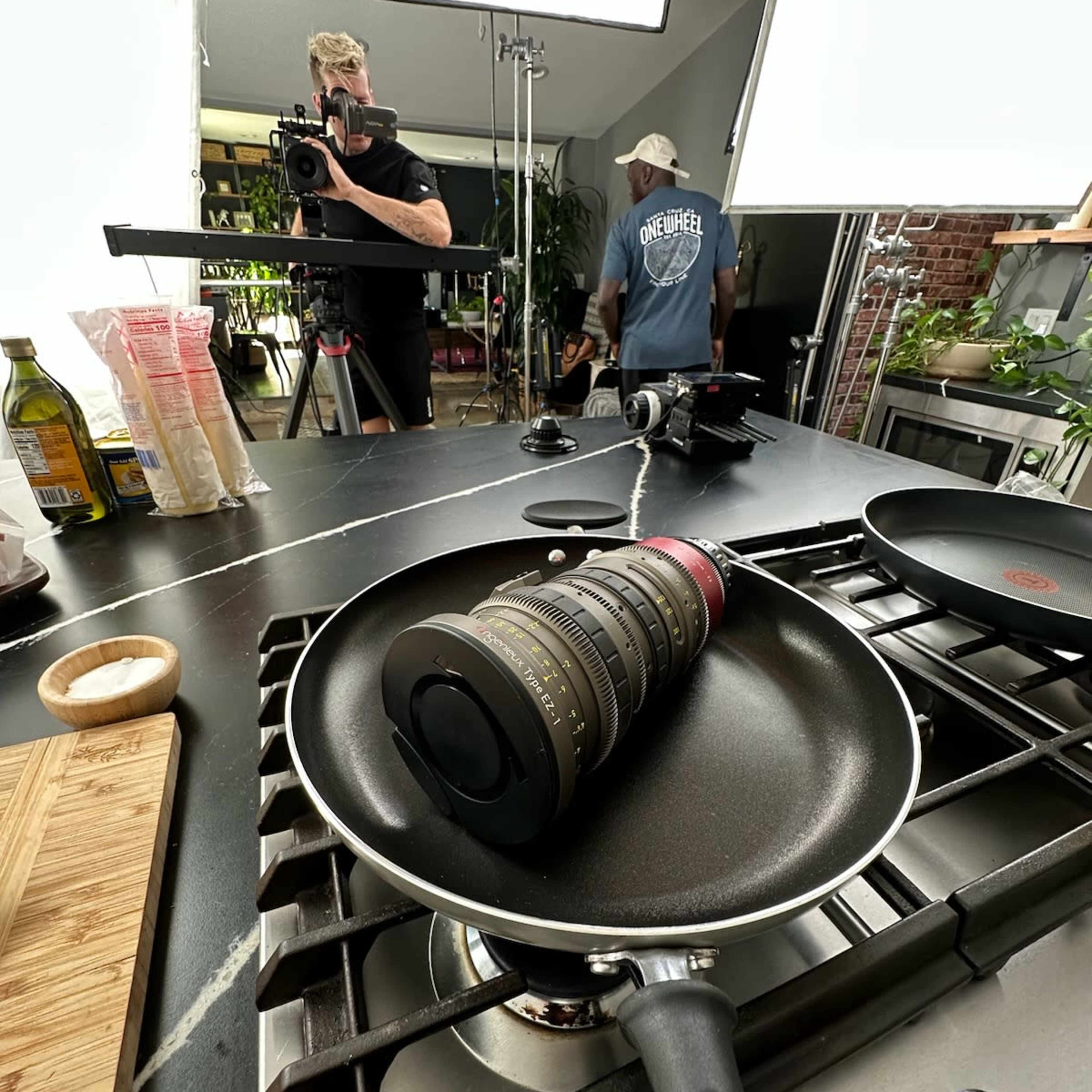 A camera is set up in a kitchen to capture a cooking scene, with a frying pan and various ingredients visible on the countertop.