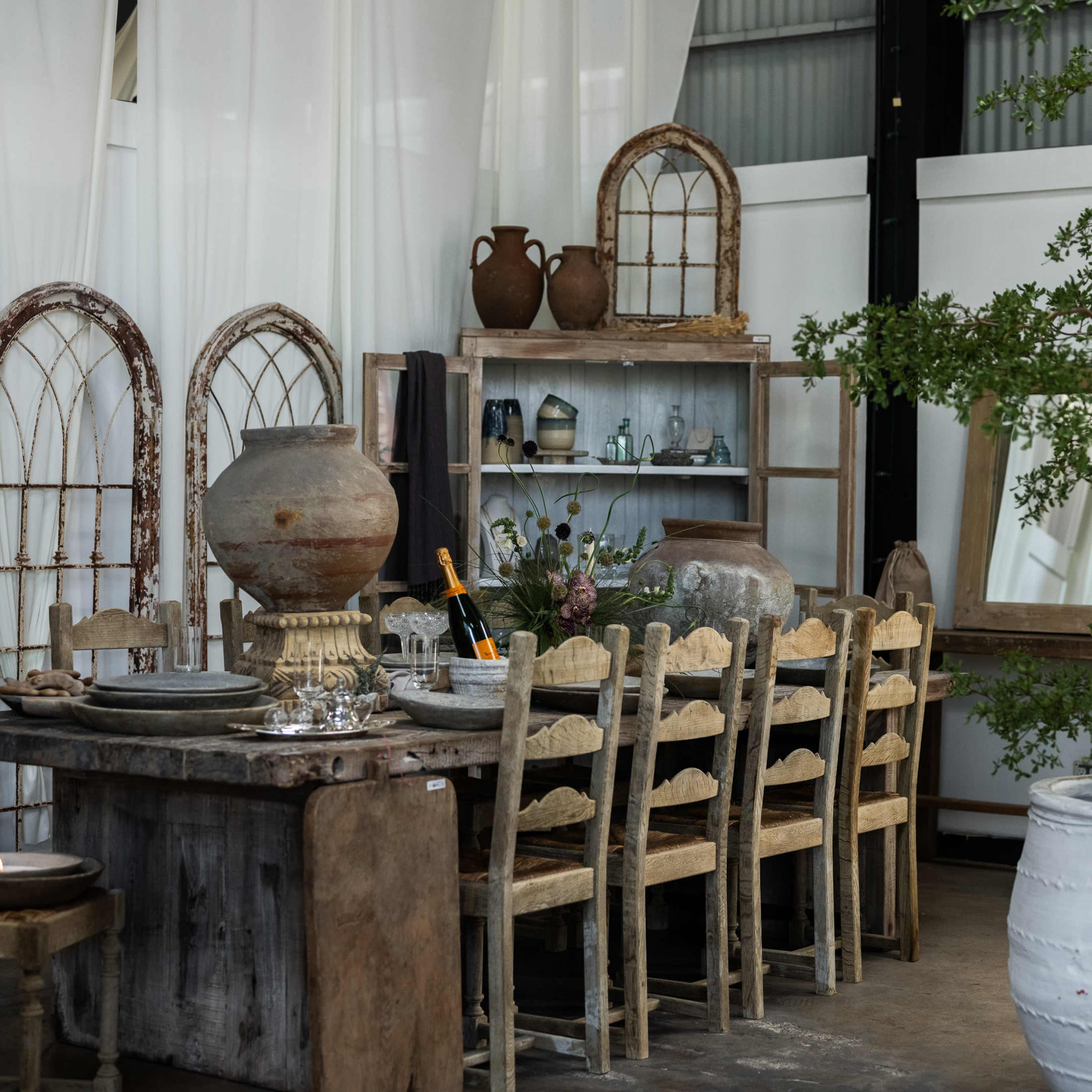 A rustic dining area with a wooden table surrounded by chairs, various pottery pieces, and decorative elements, set against a backdrop of white drapery and aged wooden accents.