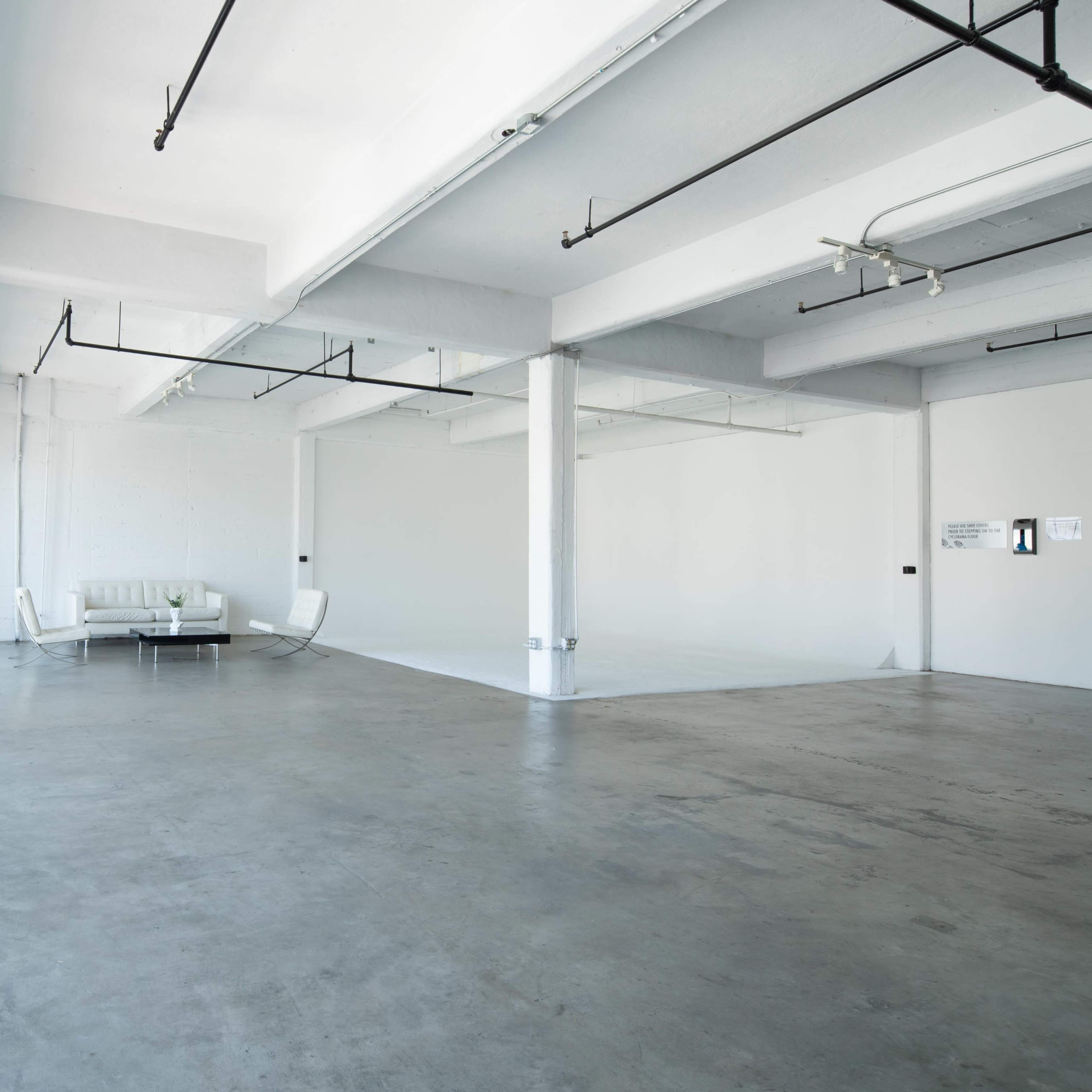 A spacious, minimalistic room with concrete flooring and large windows, featuring a white sofa and chairs.