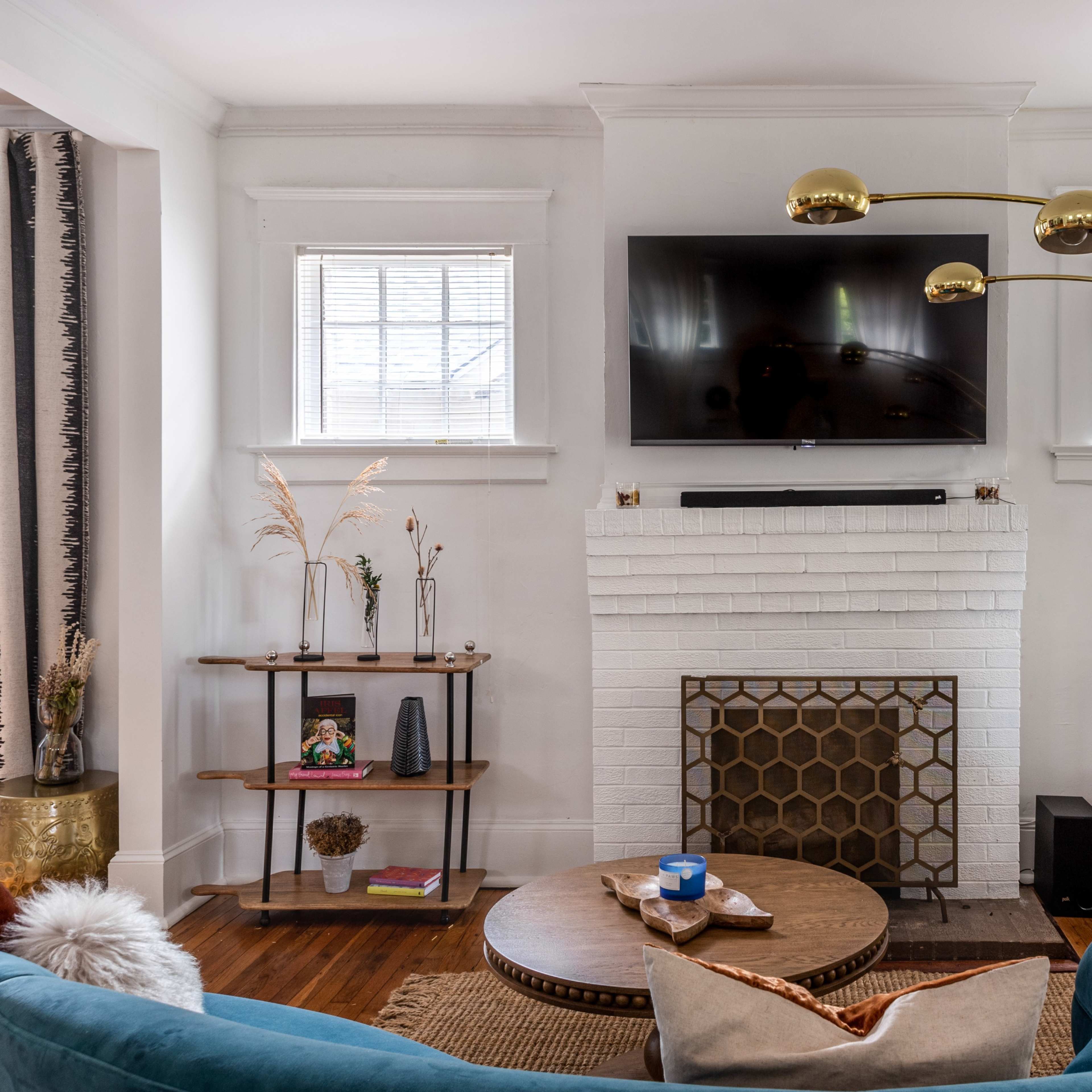 A modern living room features a teal sofa, a round coffee table, a gold-toned floor lamp, and a white-brick fireplace with a mounted television above it.