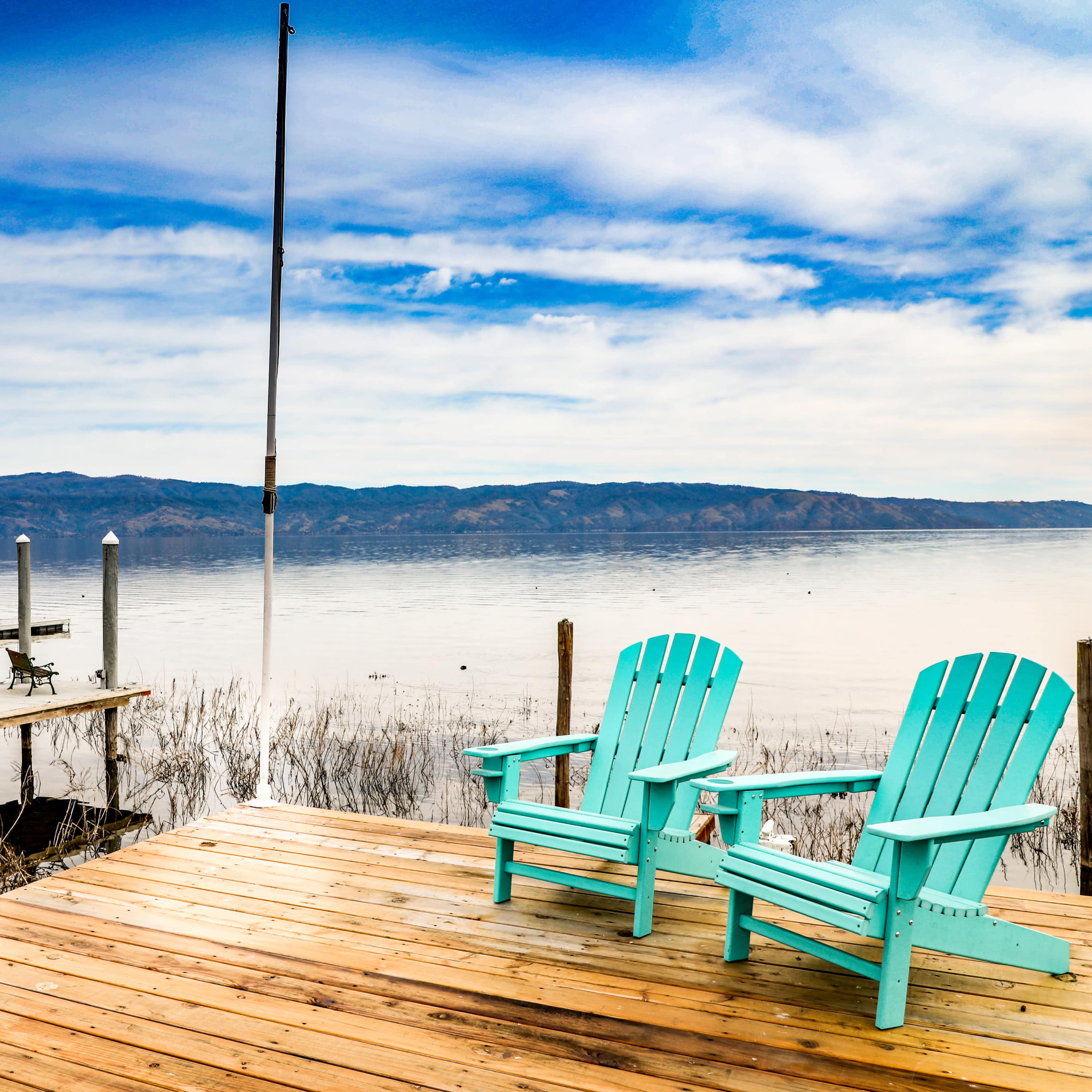 A wooden dock extends over a calm lake, featuring two turquoise Adirondack chairs facing the water and mountains in the background.