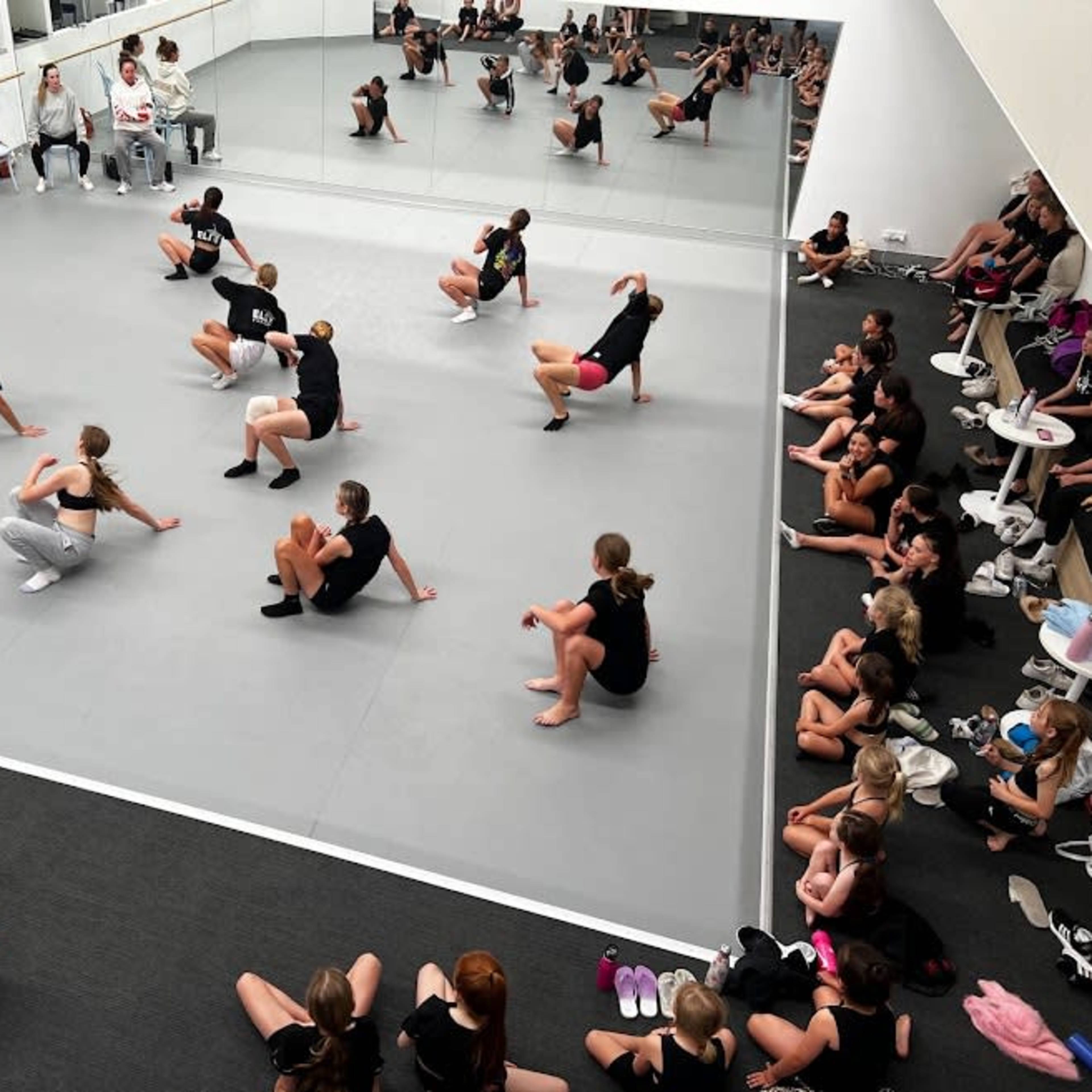 A group of dancers in black shirts practices in a mirrored studio while seated observers watch from the sidelines.