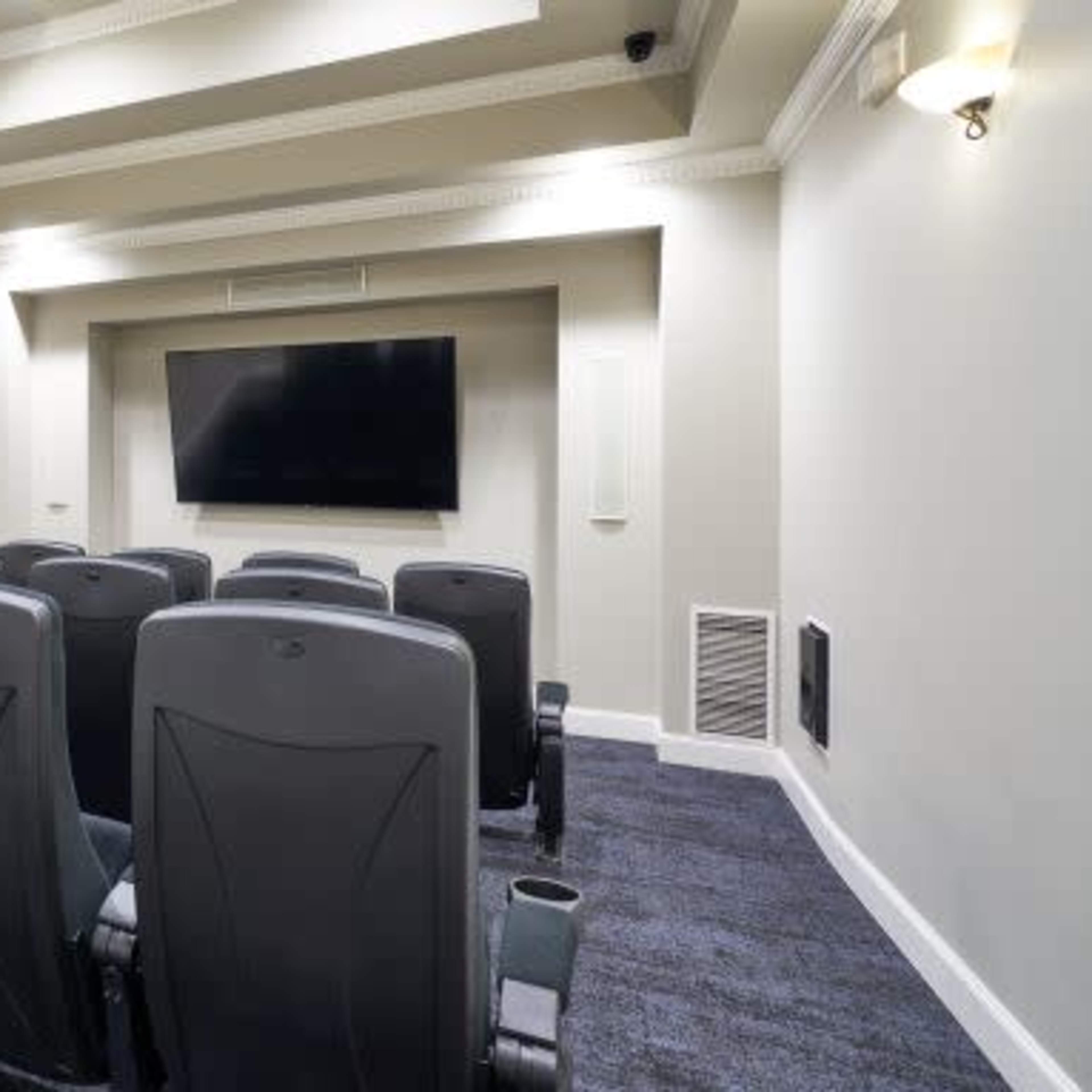 A small home theater with black seating arranged in rows facing a large flat-screen TV on a light-colored wall.
