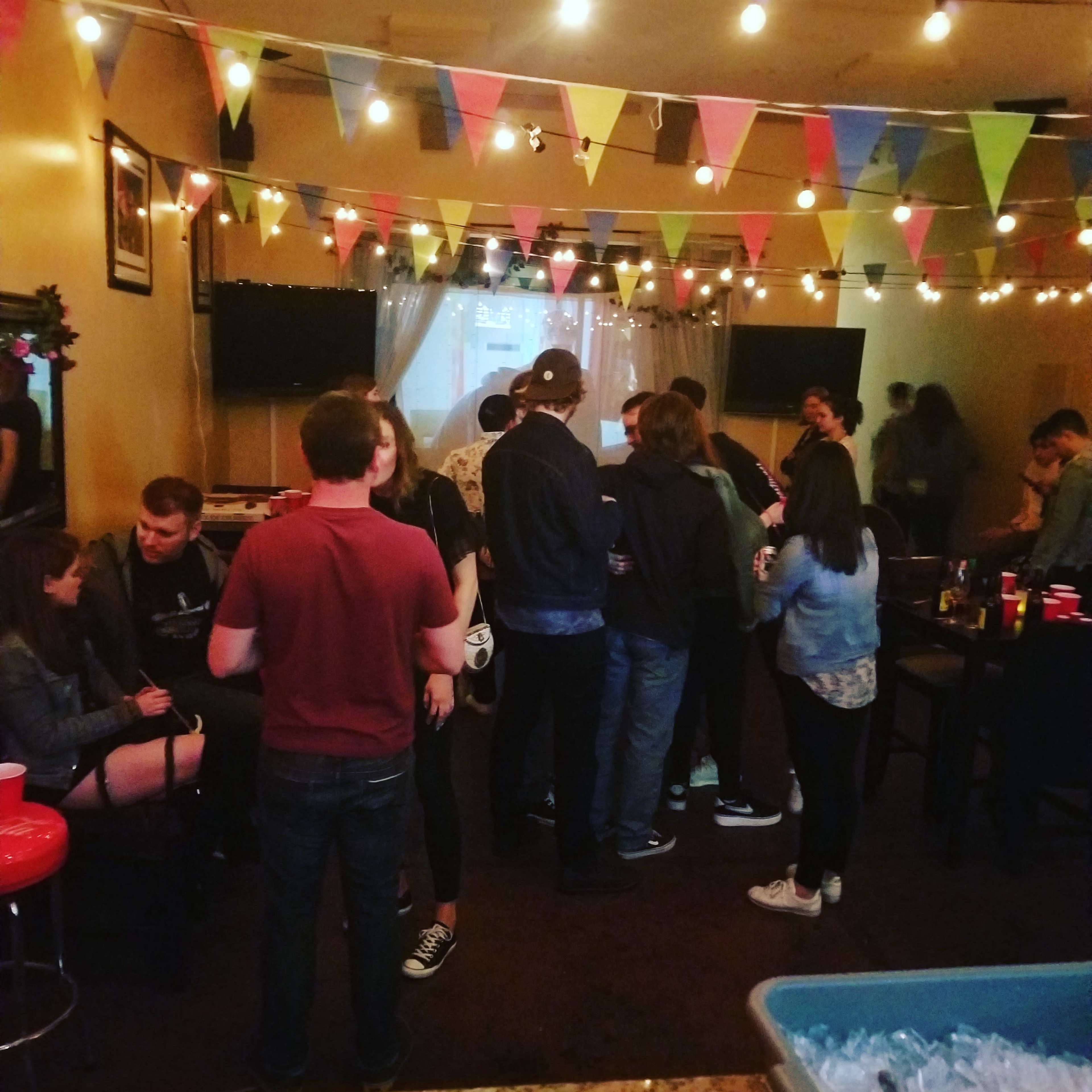 A group of people socialize at a festive gathering adorned with colorful banners and string lights.