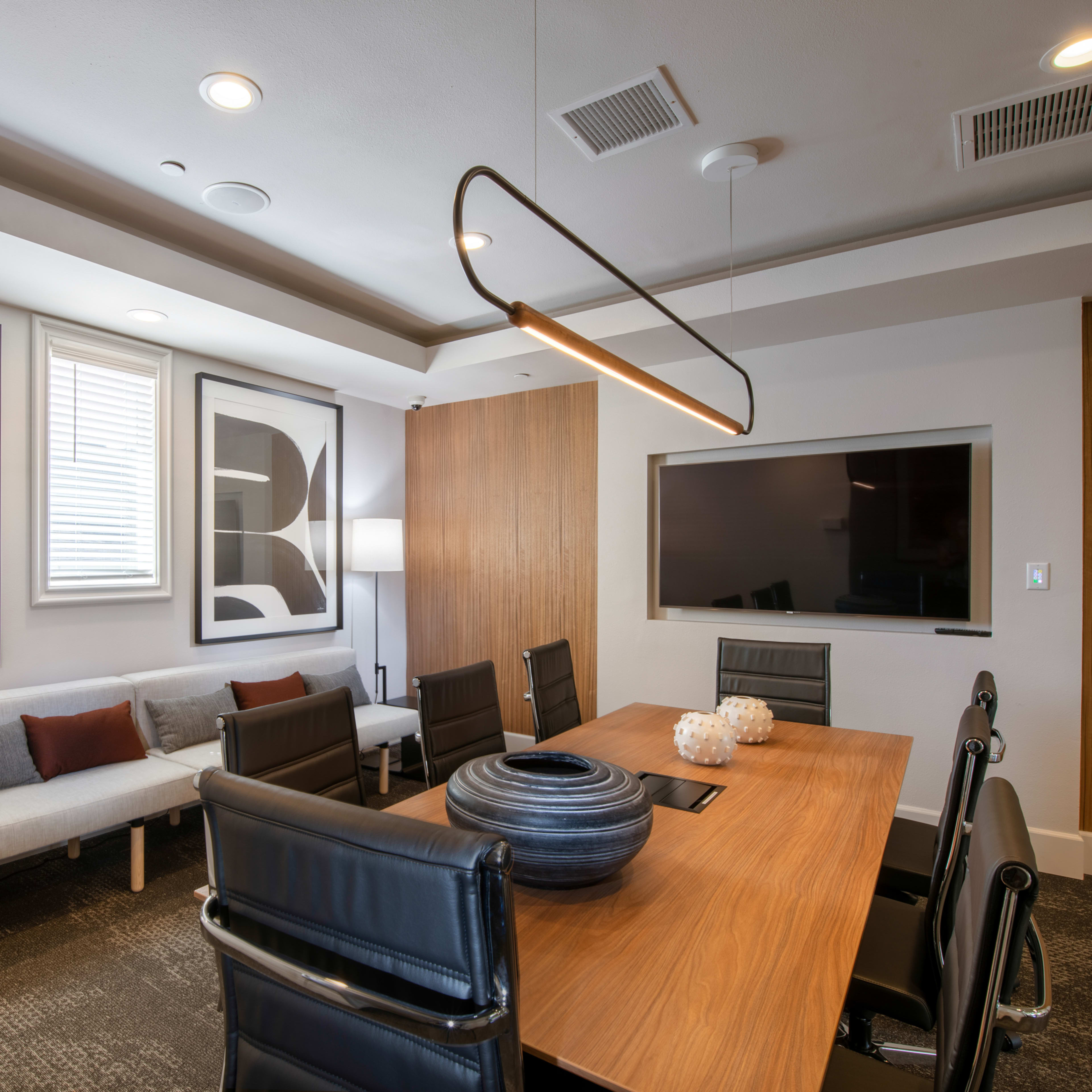 A modern conference room features a wooden table surrounded by black chairs, a light fixture overhead, and abstract artworks on the walls.