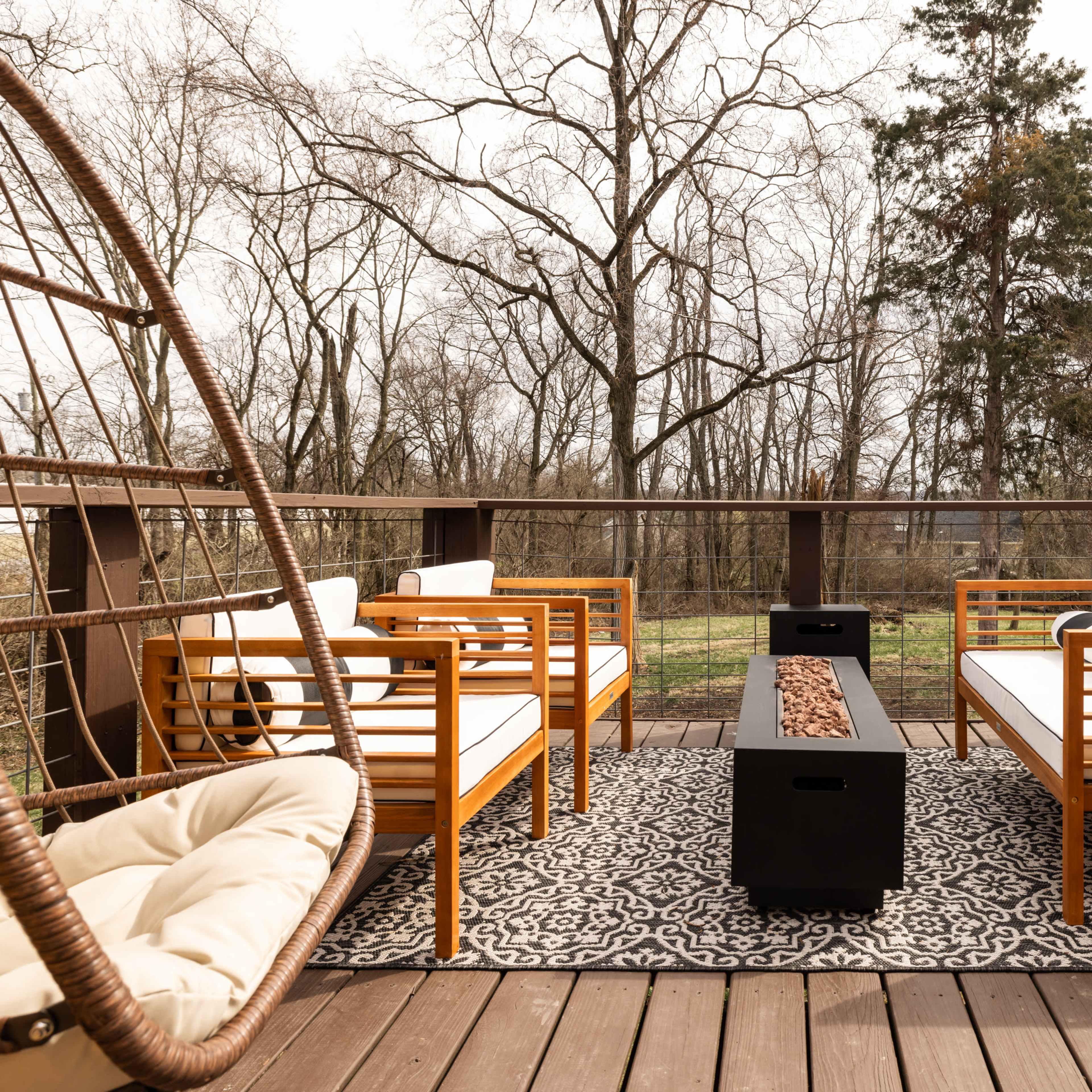 A modern outdoor seating area with wooden furniture, a central fire pit, and a rug on a wooden deck surrounded by bare trees.
