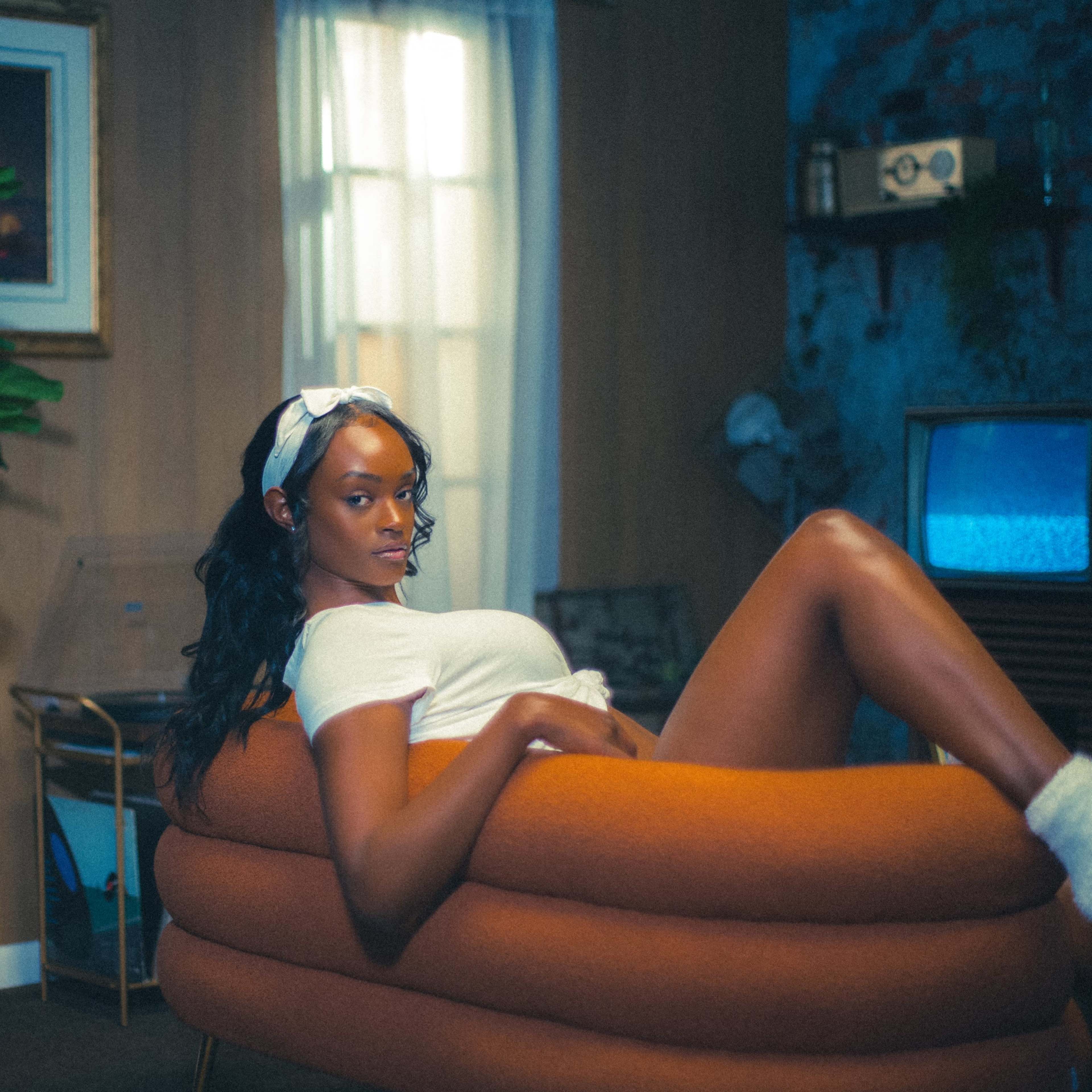 A woman sits comfortably on an orange, curved chair in a room with vintage decor, including plants and a retro television.