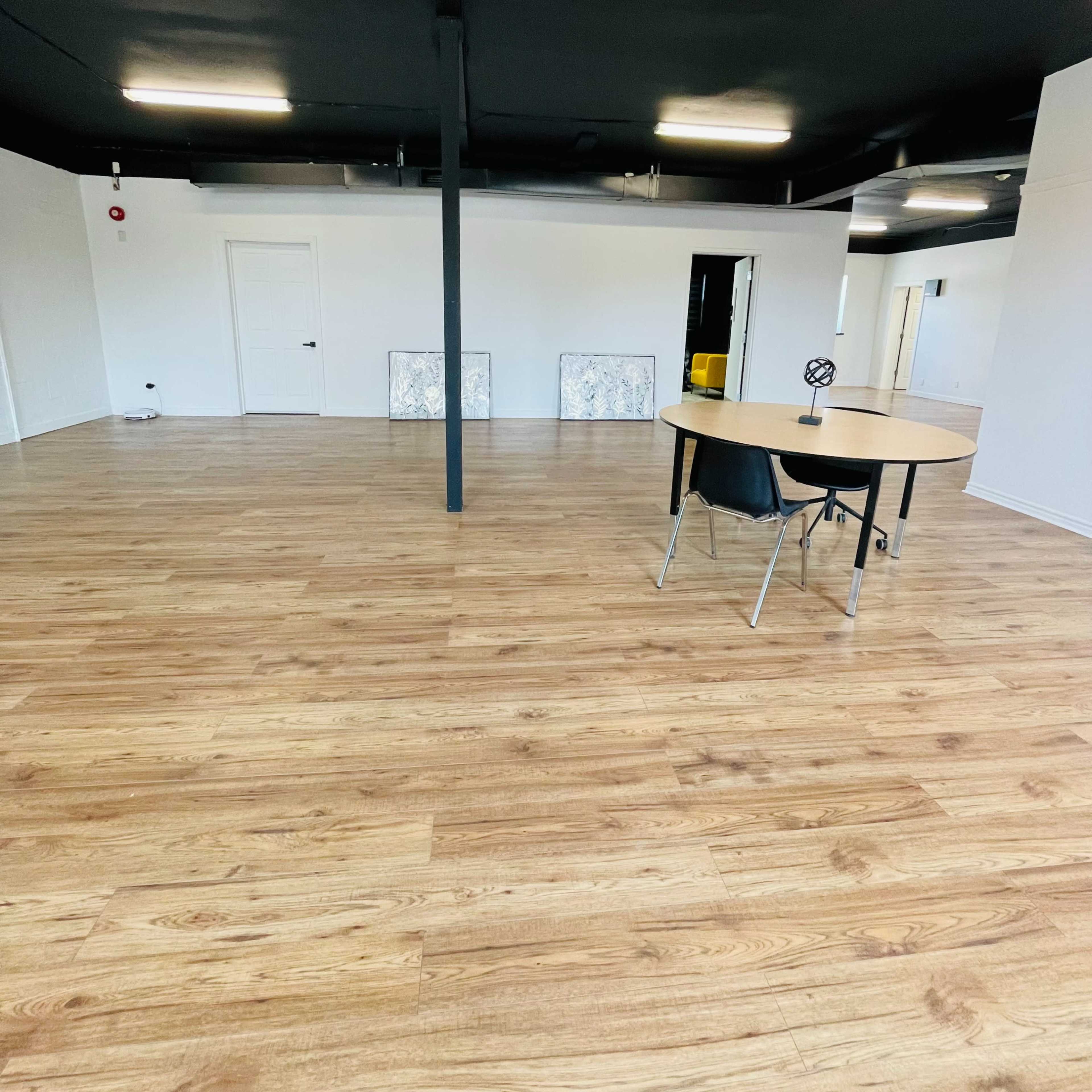 A spacious, bare room with wooden flooring, featuring a round table and chair in the foreground, and white walls with a few doors and two framed artworks leaning against the wall in the background.